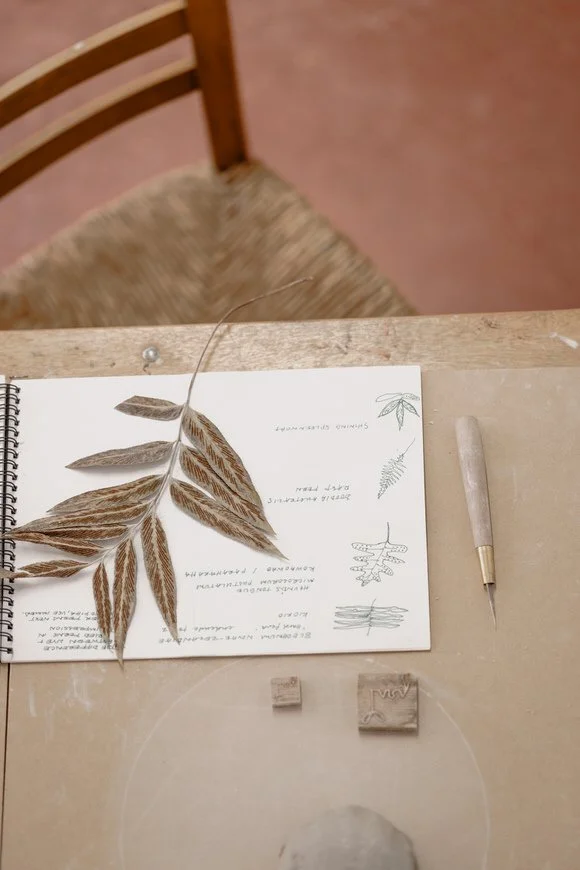 Open notebook with a pressed leaf and botanical sketches on a wooden table, next to a carving tool and small clay stamps.