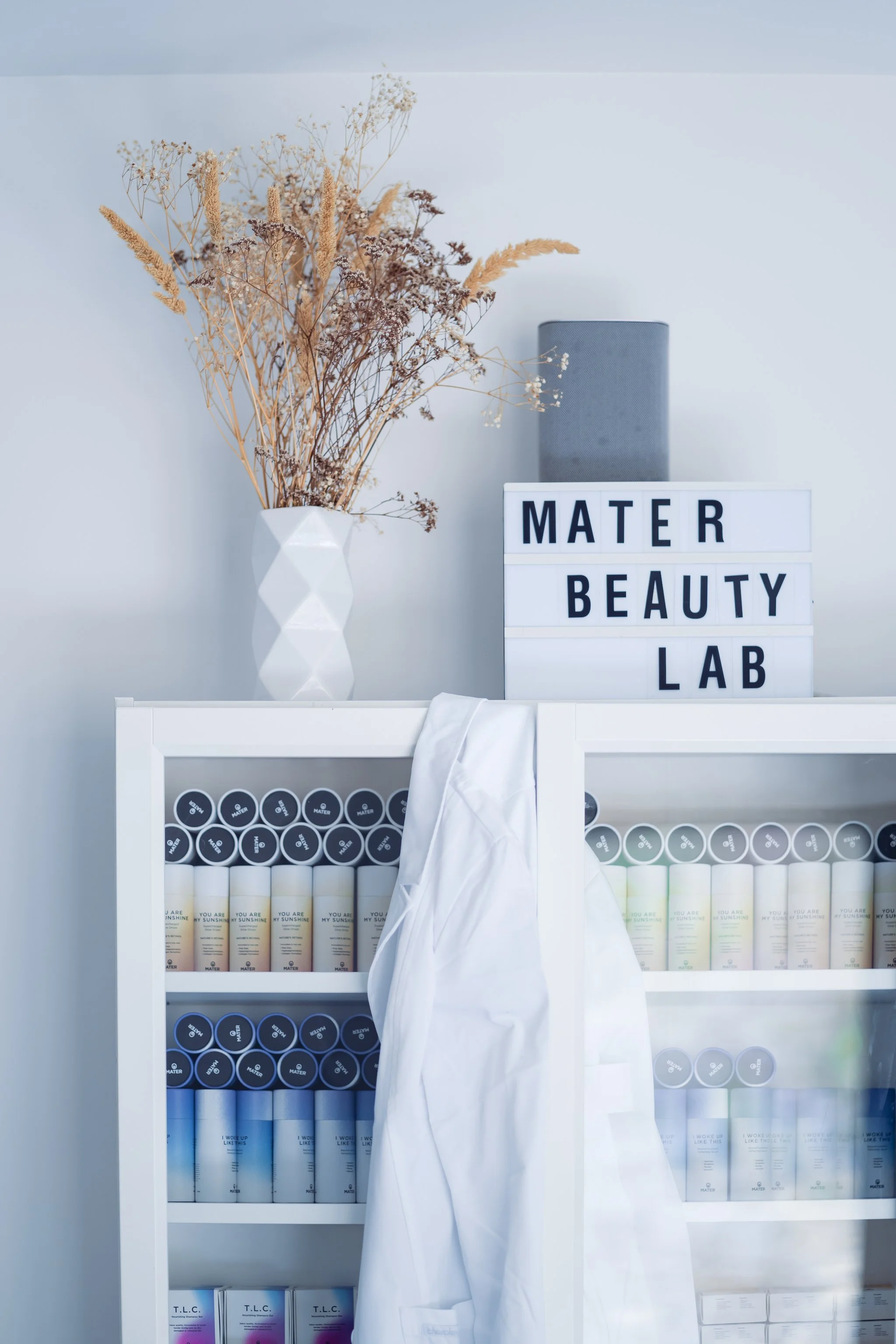 Shelf with beauty products, a white lab coat, and a vase with dried flowers; sign reads 'Mater Beauty Lab.'