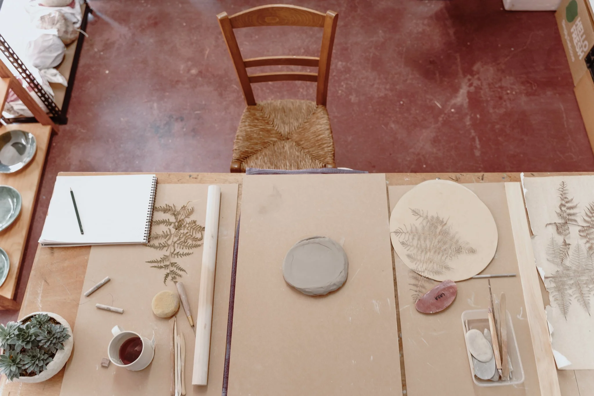 Top view of a pottery workspace with a wooden chair. The table has clay, a rolling pin, a notebook with a pencil, and artistic tools. Fern leaves are being used for imprinting designs. A potted plant and a coffee cup are also visible.