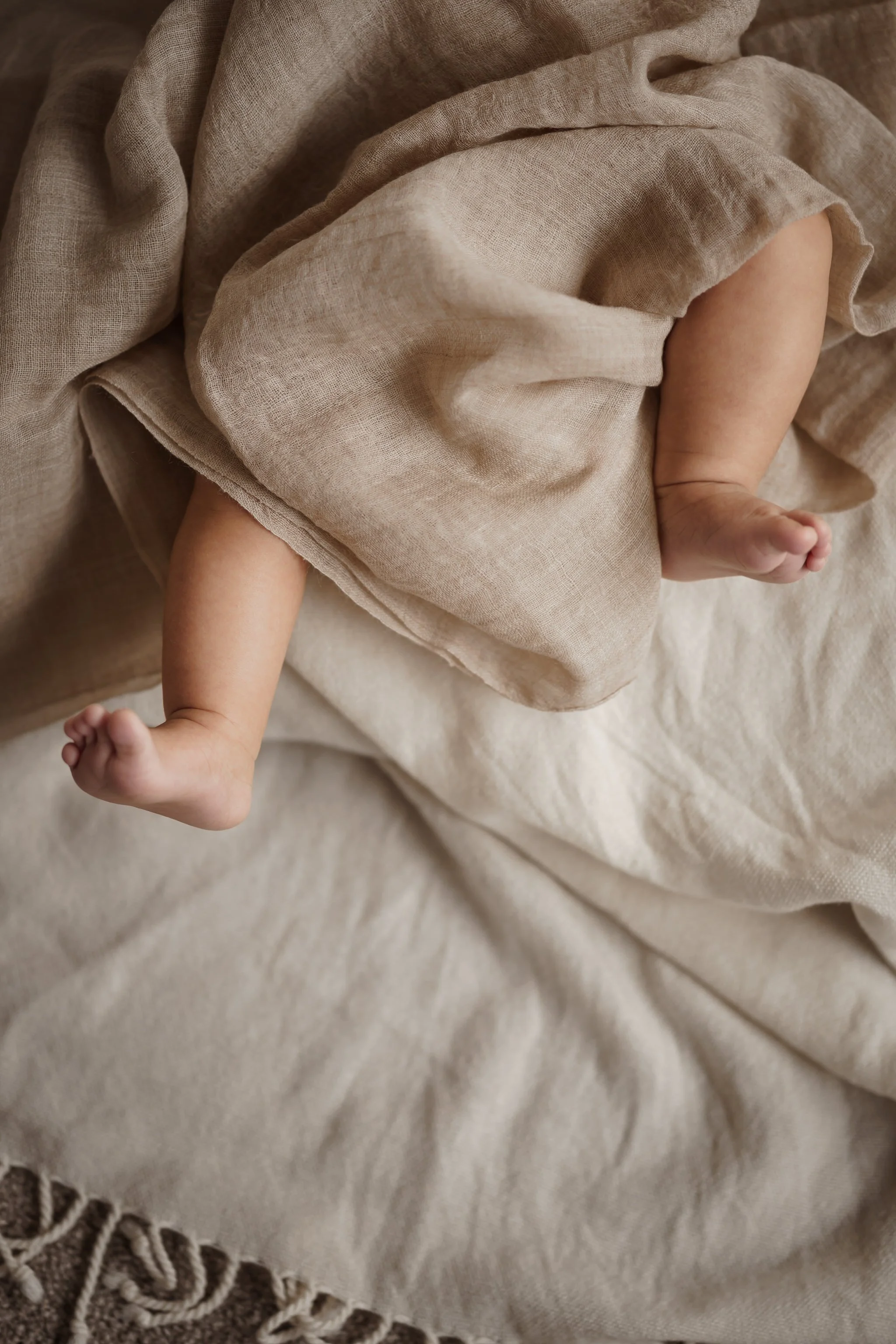 Baby feet peeking from under a beige blanket on a soft fabric surface.