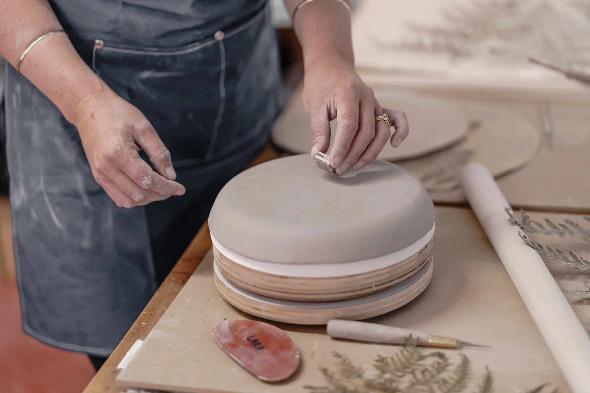 Person working on a clay piece in pottery studio, smoothing surface with tool, surrounded by rolling pin and clay tools.