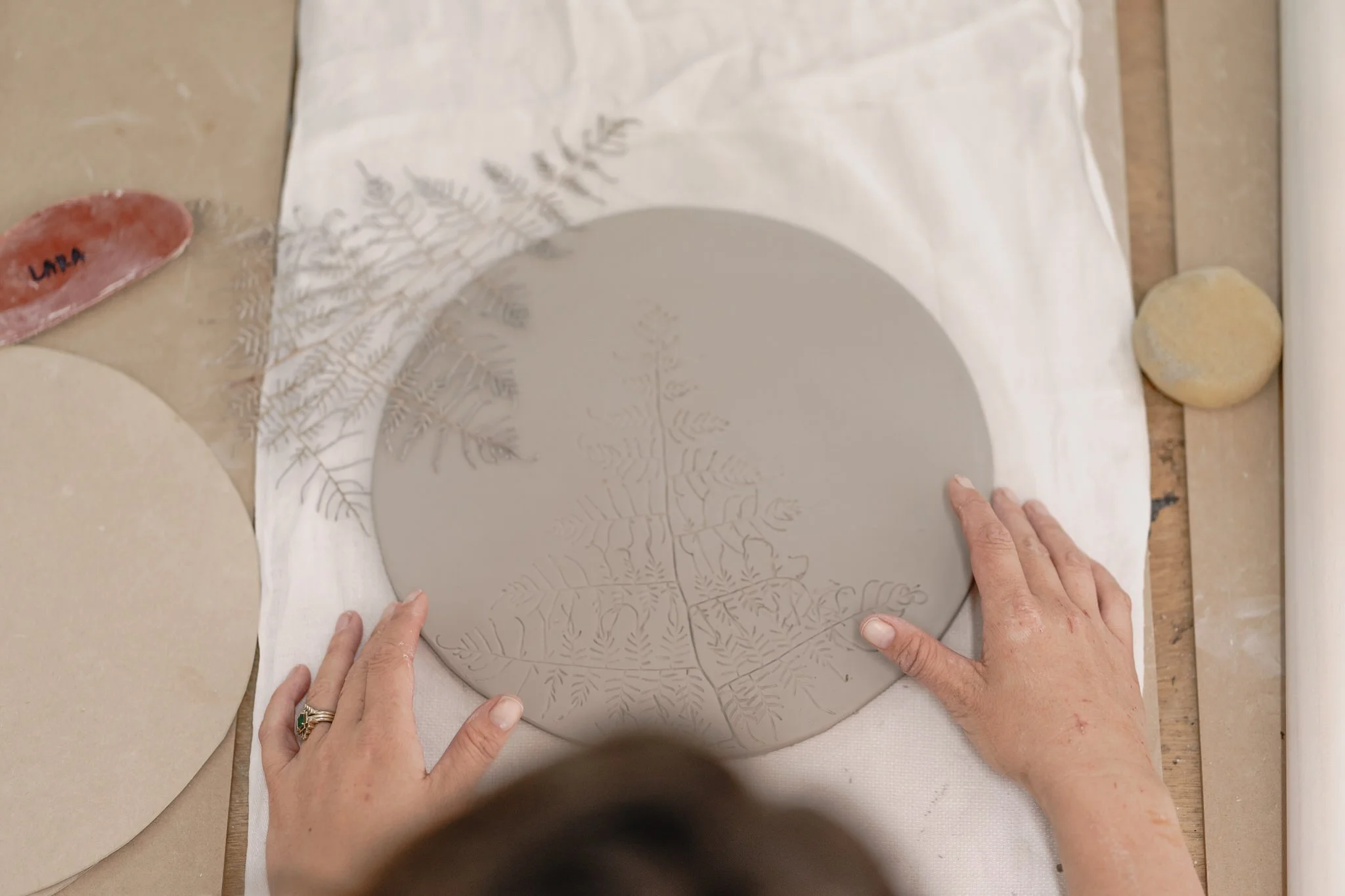Hands pressing ferns onto a circular clay slab for texture patterning, on a worktable.
