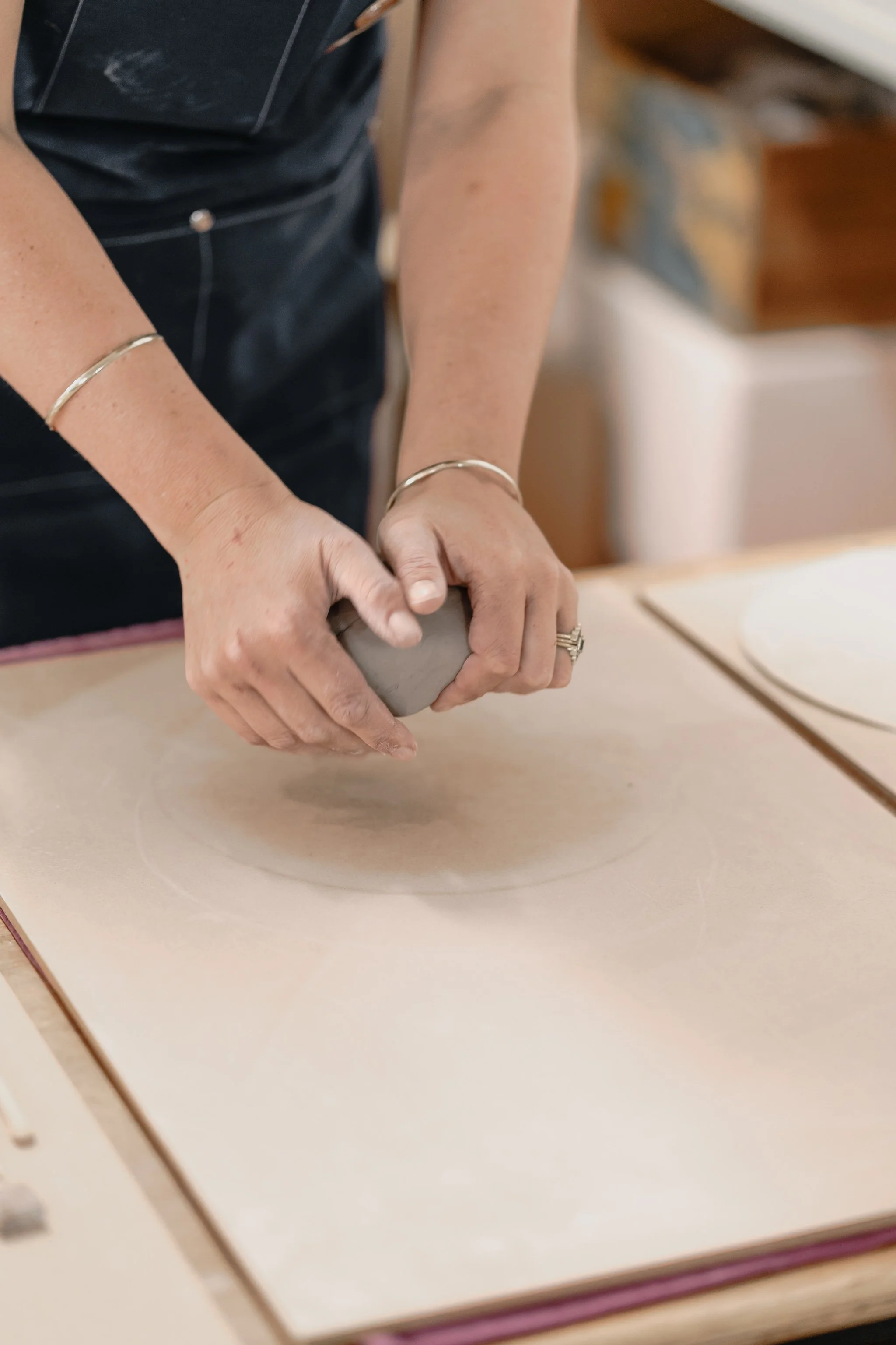 Person kneading clay on a workbench, wearing a dark apron and bracelets.