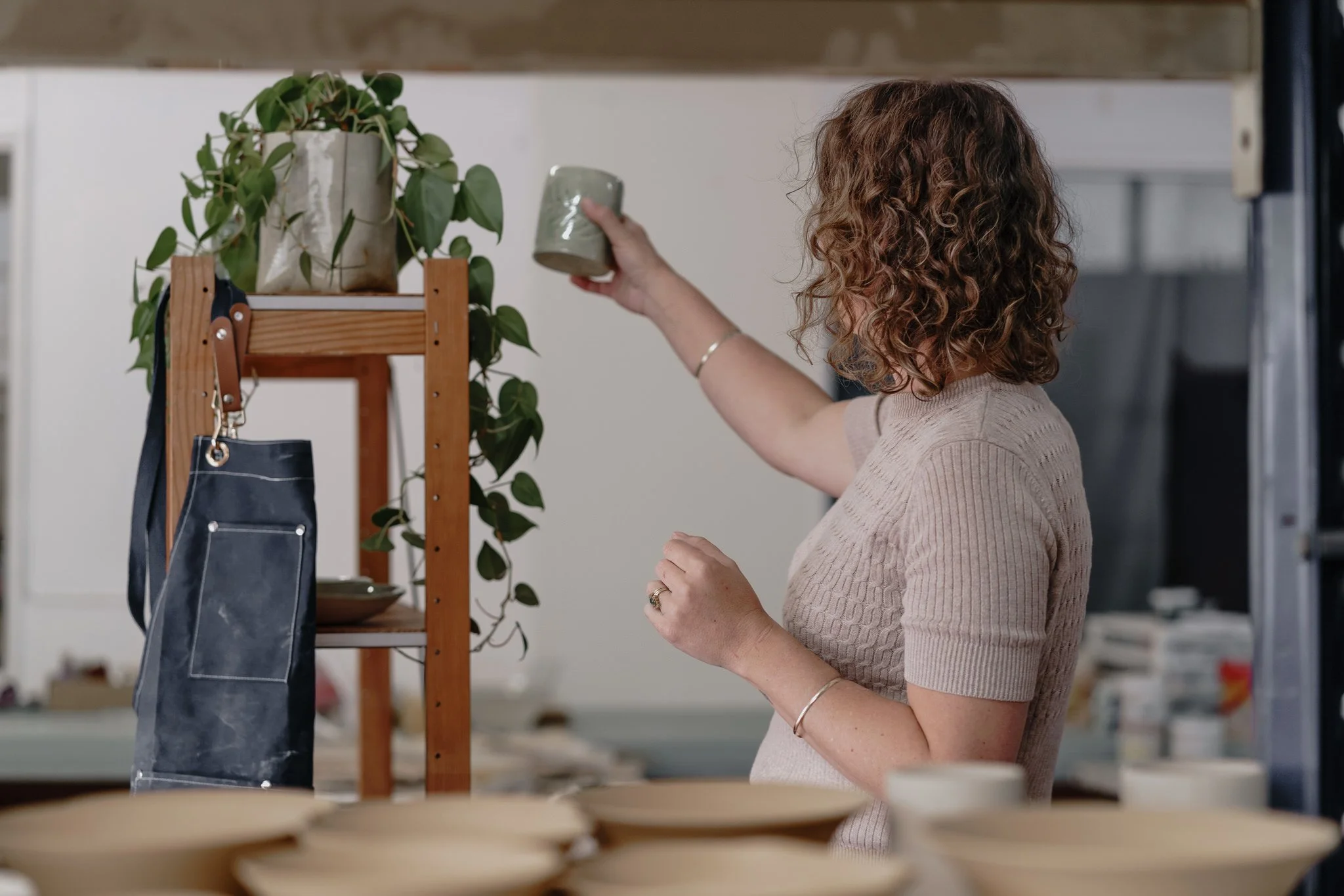Person in a pottery studio holding a ceramic cup next to shelves with plants and a hanging apron.