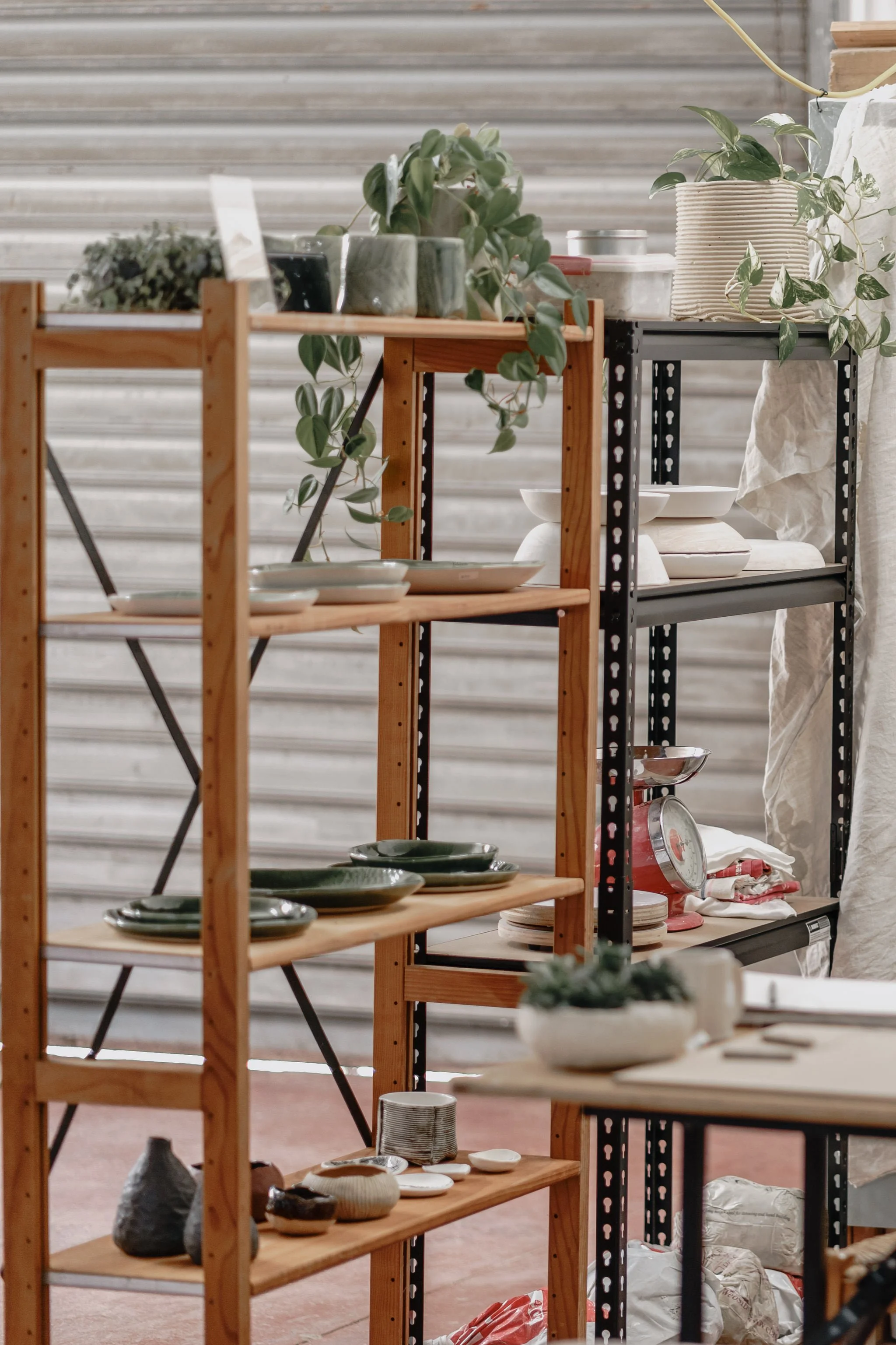 Wooden and metal shelves with ceramic plates, bowls, potted plants, and a weighing scale in a rustic setting.