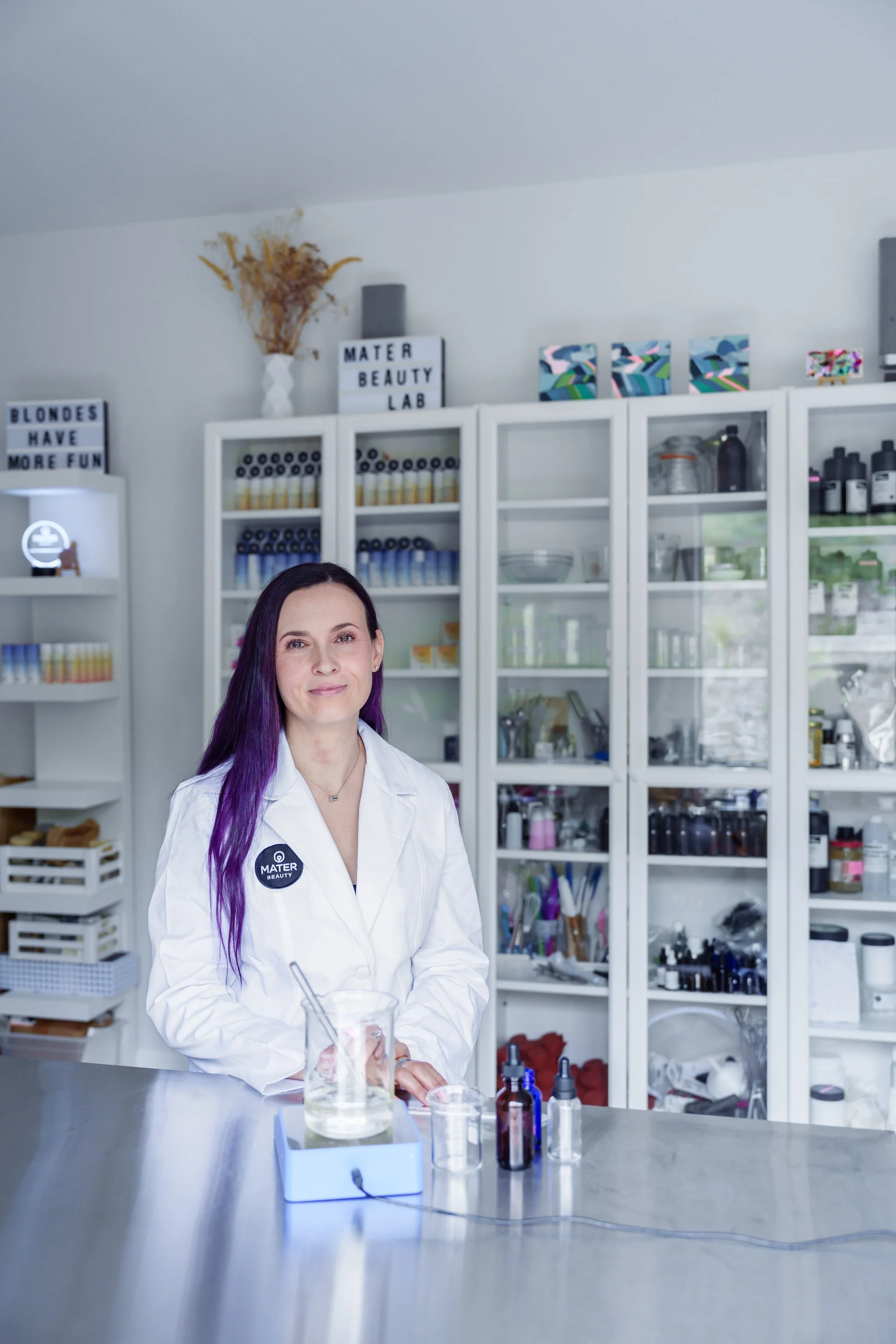 Woman in a lab coat standing in a beauty lab with shelves of bottles and equipment.