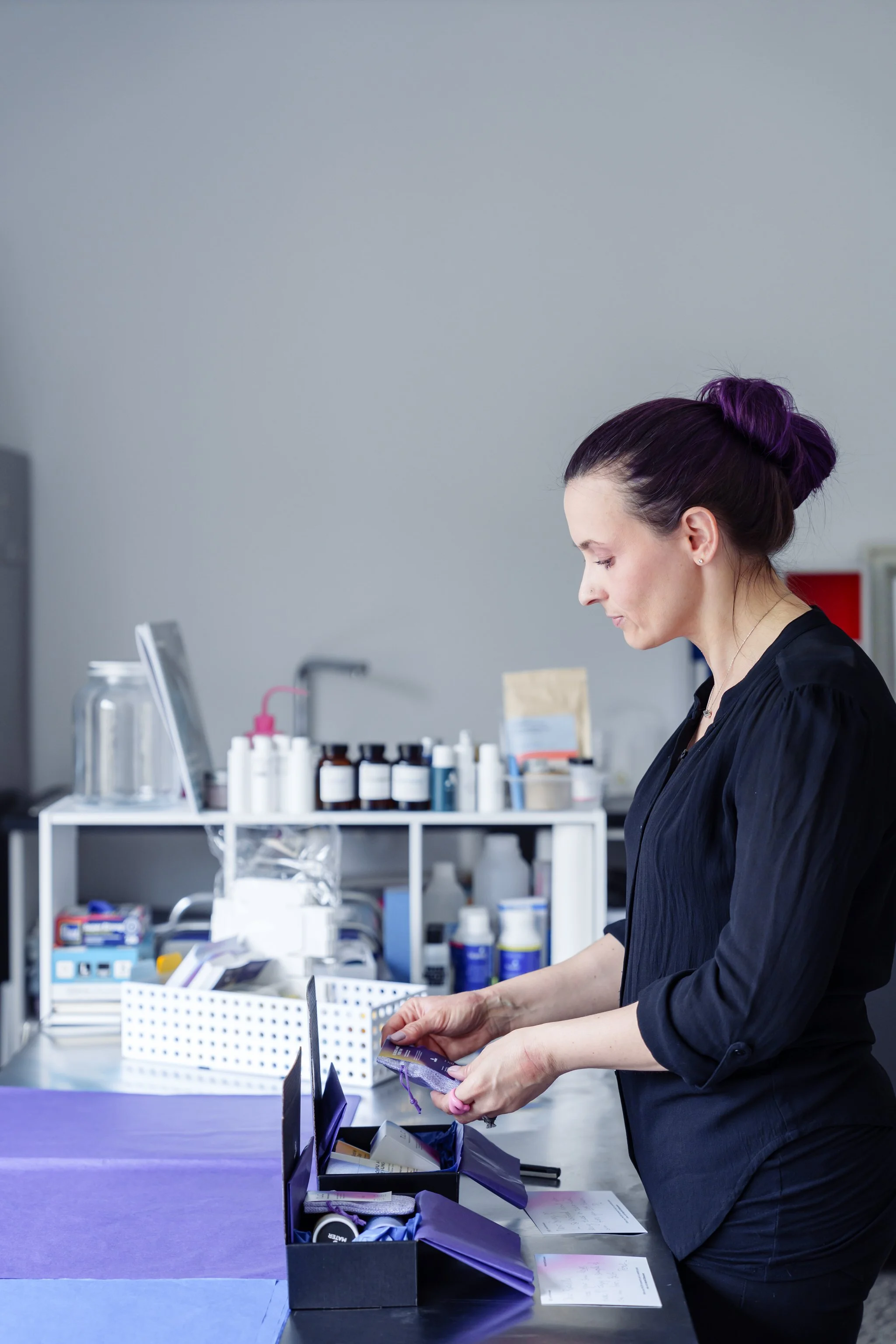 Person with purple hair organizing cosmetics on a table in a lab setting, with bottles and jars on shelves in the background.