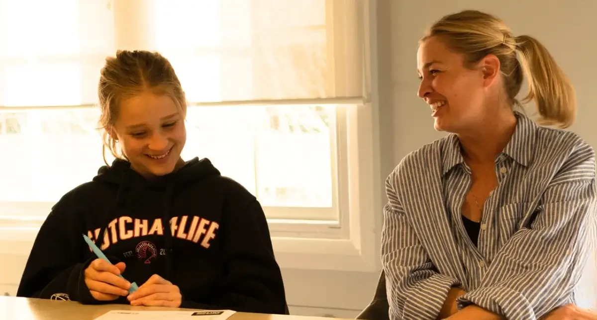 A female child in a black hoodie writes at a table as her smiling mother sits beside her in a bright room, sharing a warm, supportive moment.