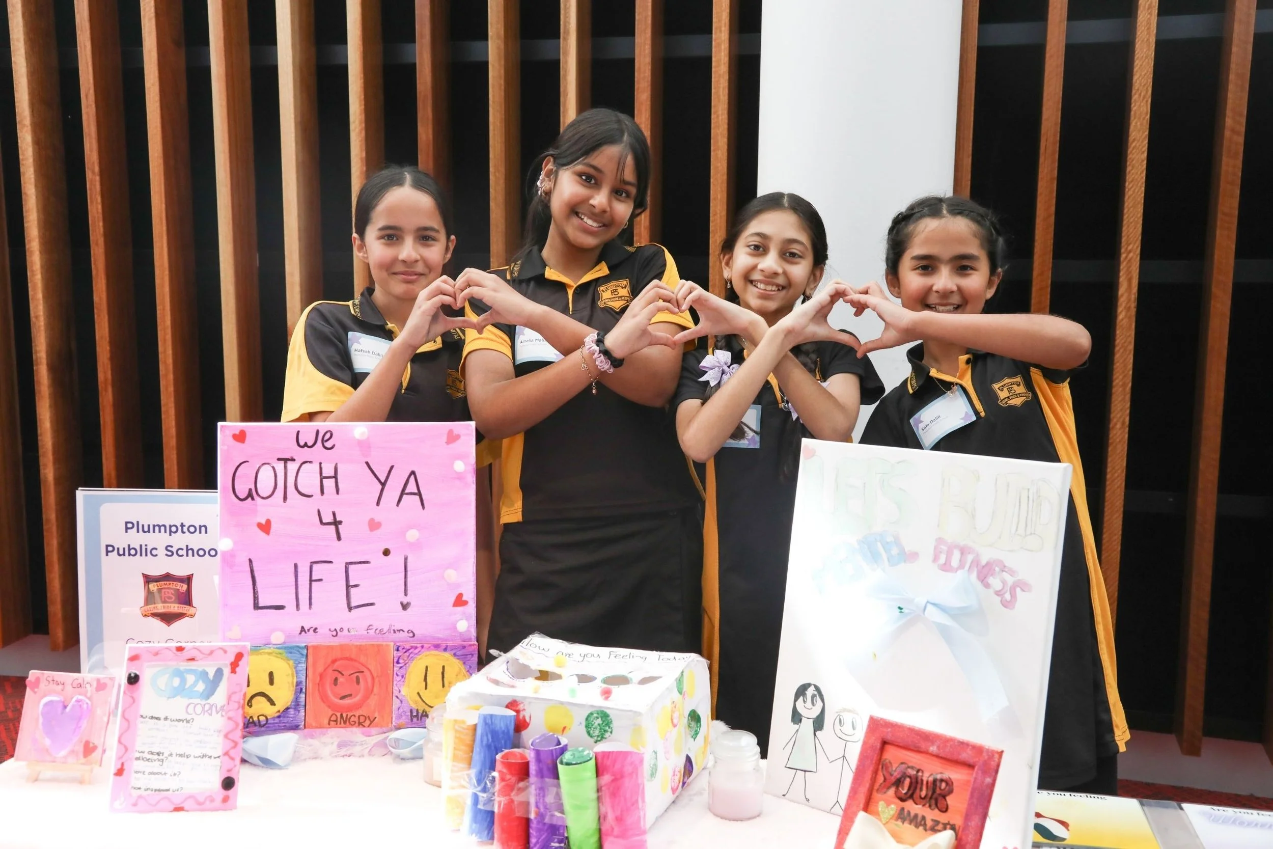 Four female primary school students standing next to each other and smiling as they make joint heart shapes with their hands