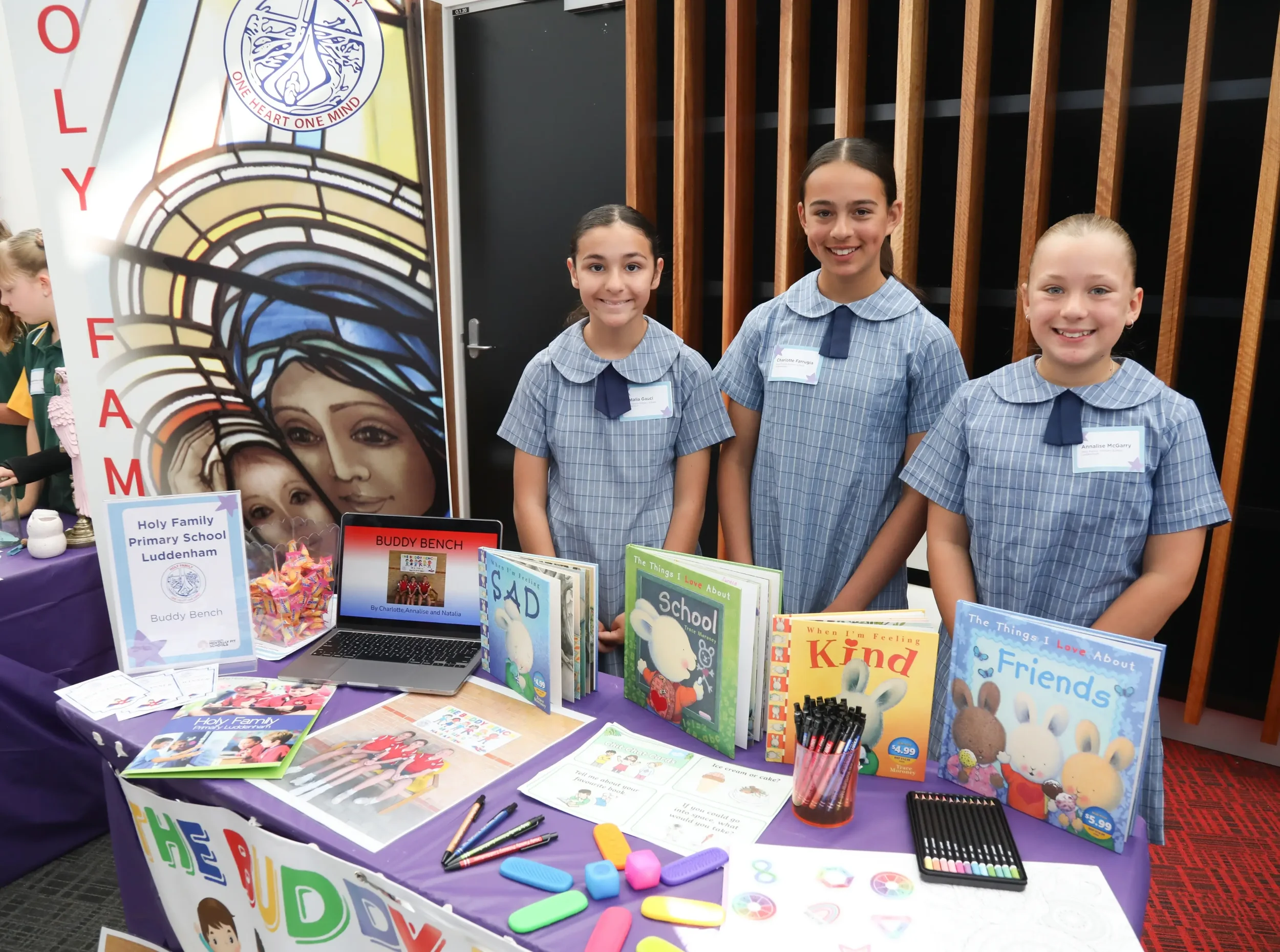 Three students from Holy Family Luddenham school stand at their display at Gotcha4Life's Mental Fitness Leaders of Tomorrow event, with books and writing materials on the table top.