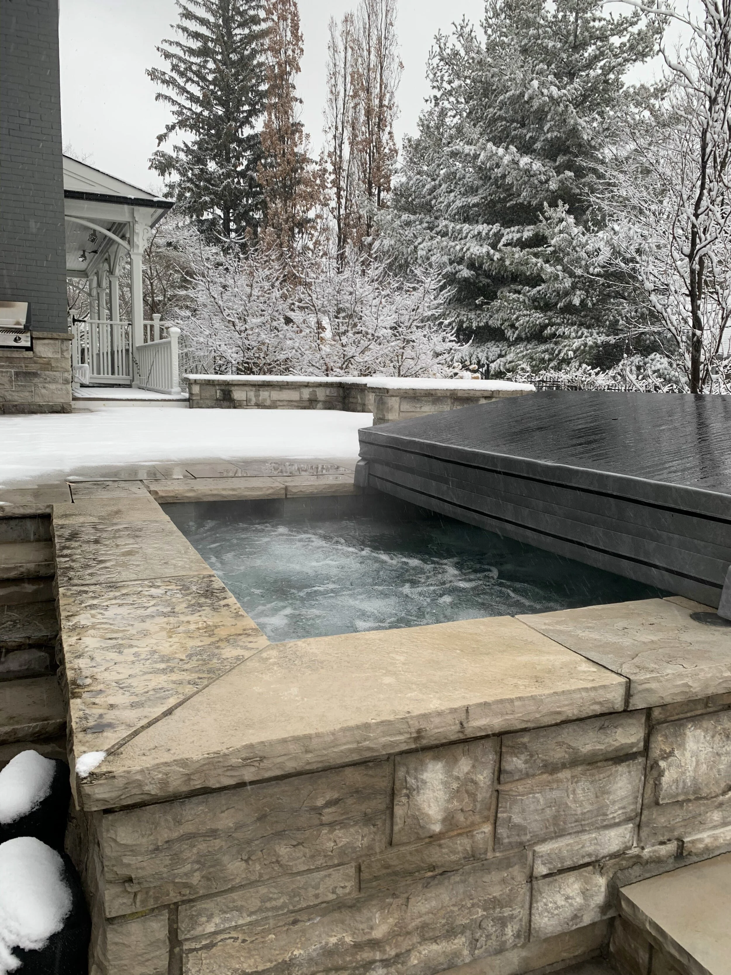 A backyard with a stone patio, a hot tub covered with a dark cover, and a snow-covered deck, with snow-covered trees and a house in the background.