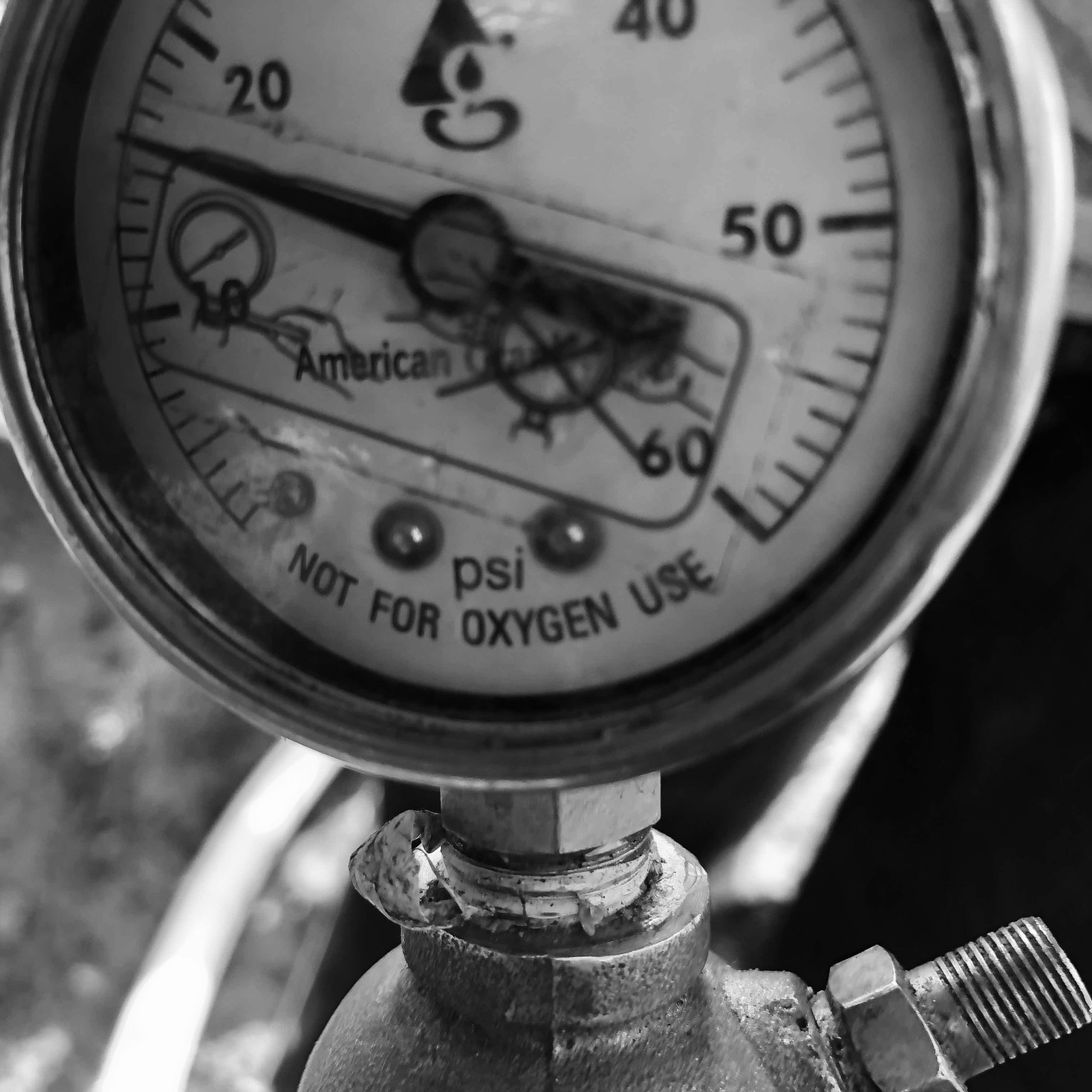 Close-up black and white photo of a pressure gauge with the words 'American' and 'Linde' visible, attached to pipes or machinery.