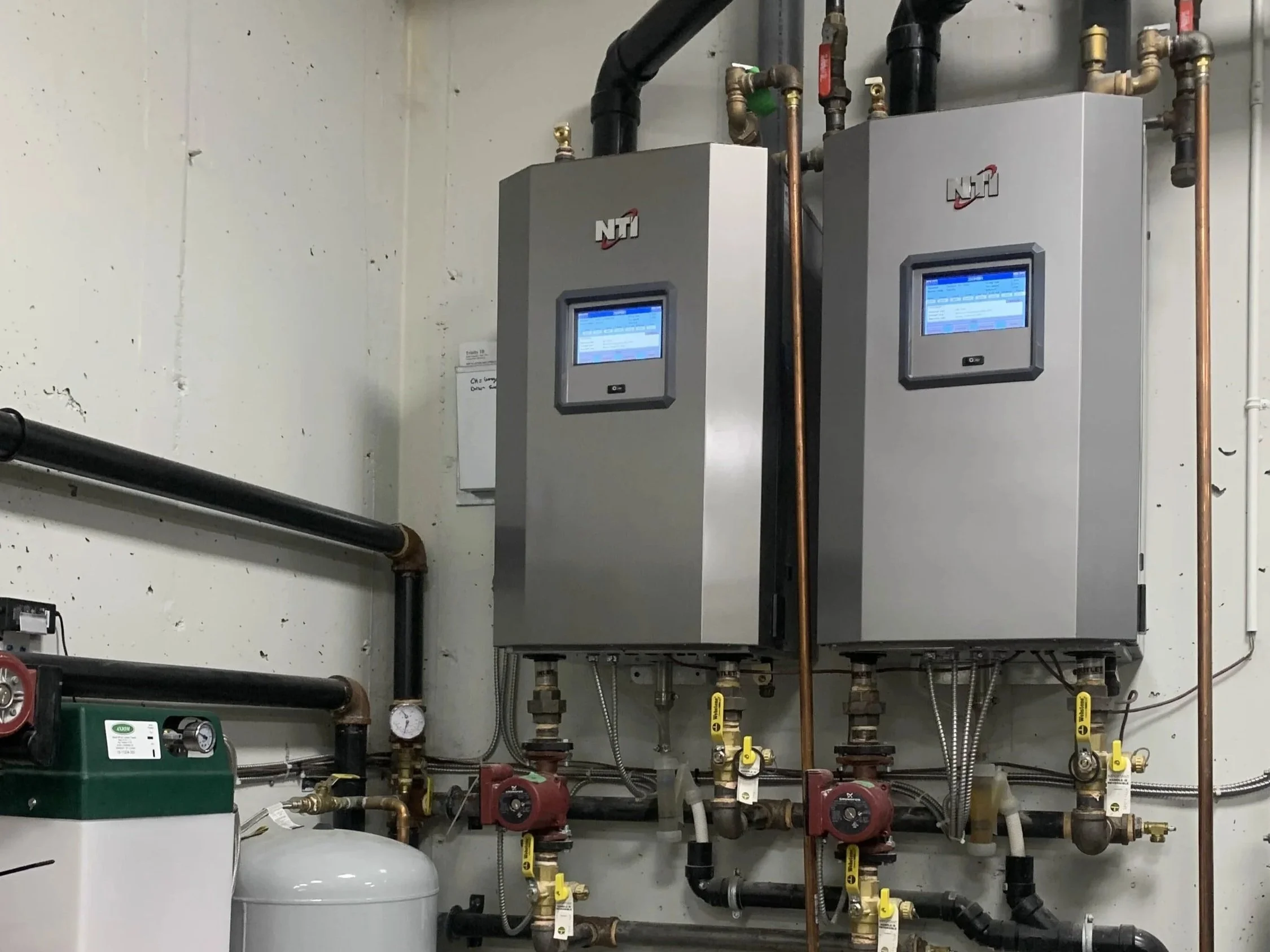 Internal view of a mechanical room with two modern electronic control units or panels branded NTI, connected to pipes, valves, and gauges for plumbing or heating system management.
