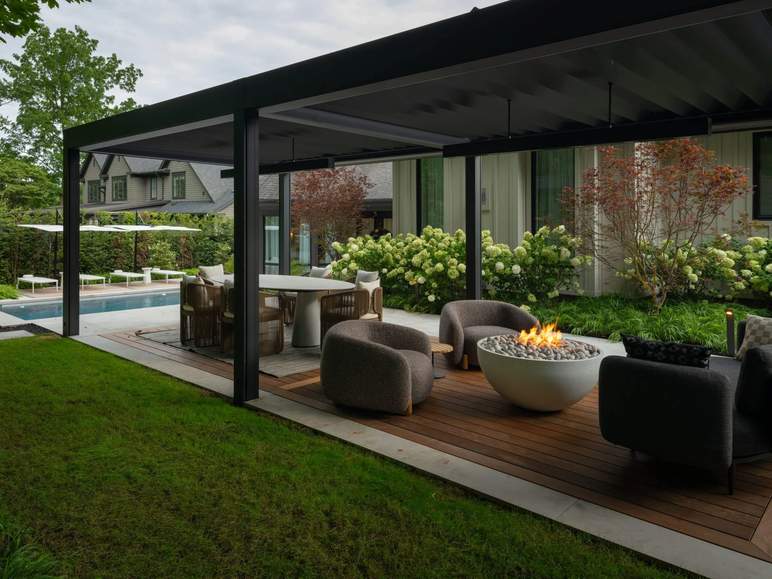Custom fire pit set within an outdoor patio area with modern chairs and lush green plants. Part of a patio house with large glass windows.