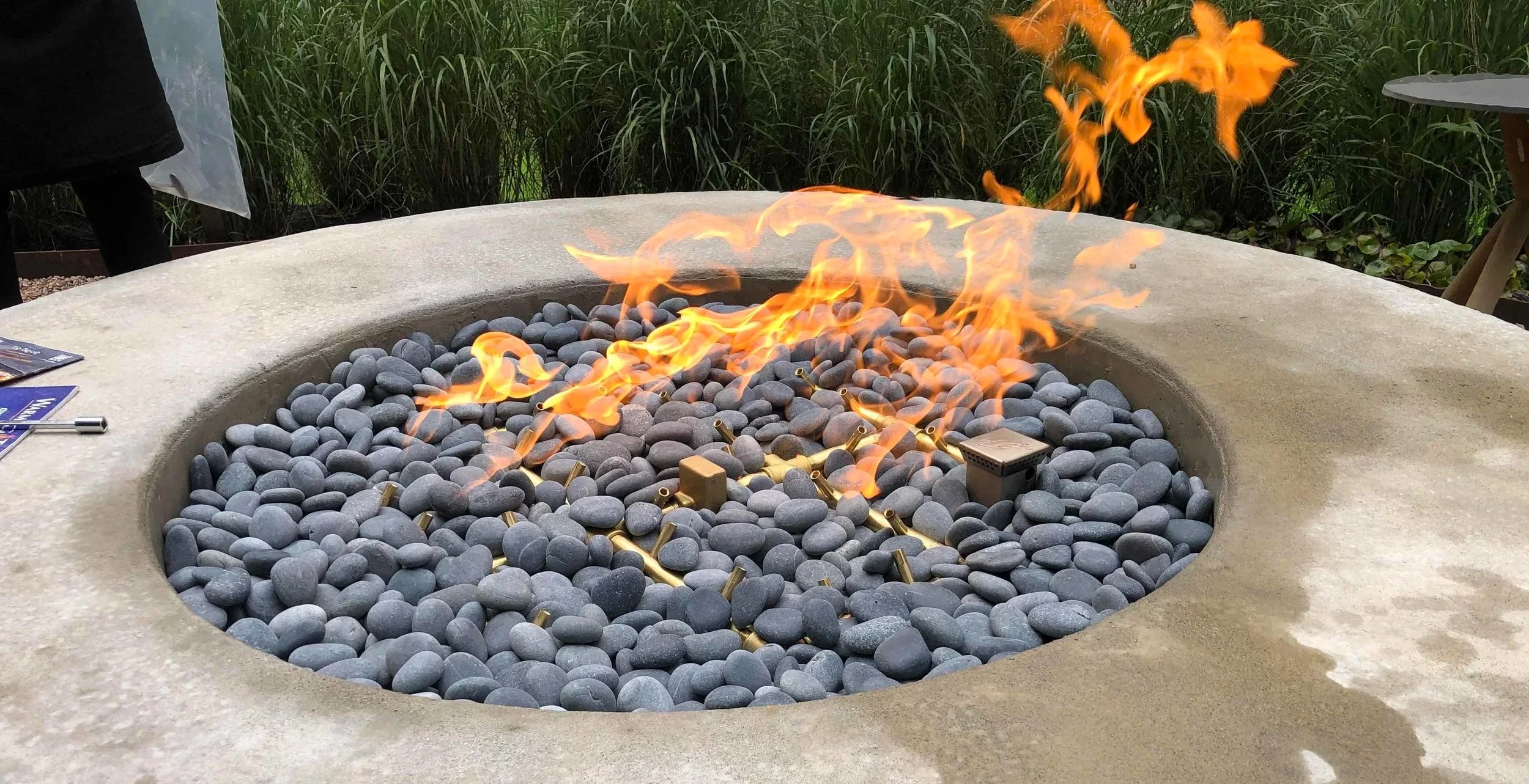 A fire pit filled with smooth gray stones and lit with flames, surrounded by a concrete or stone border and greenery in the background.