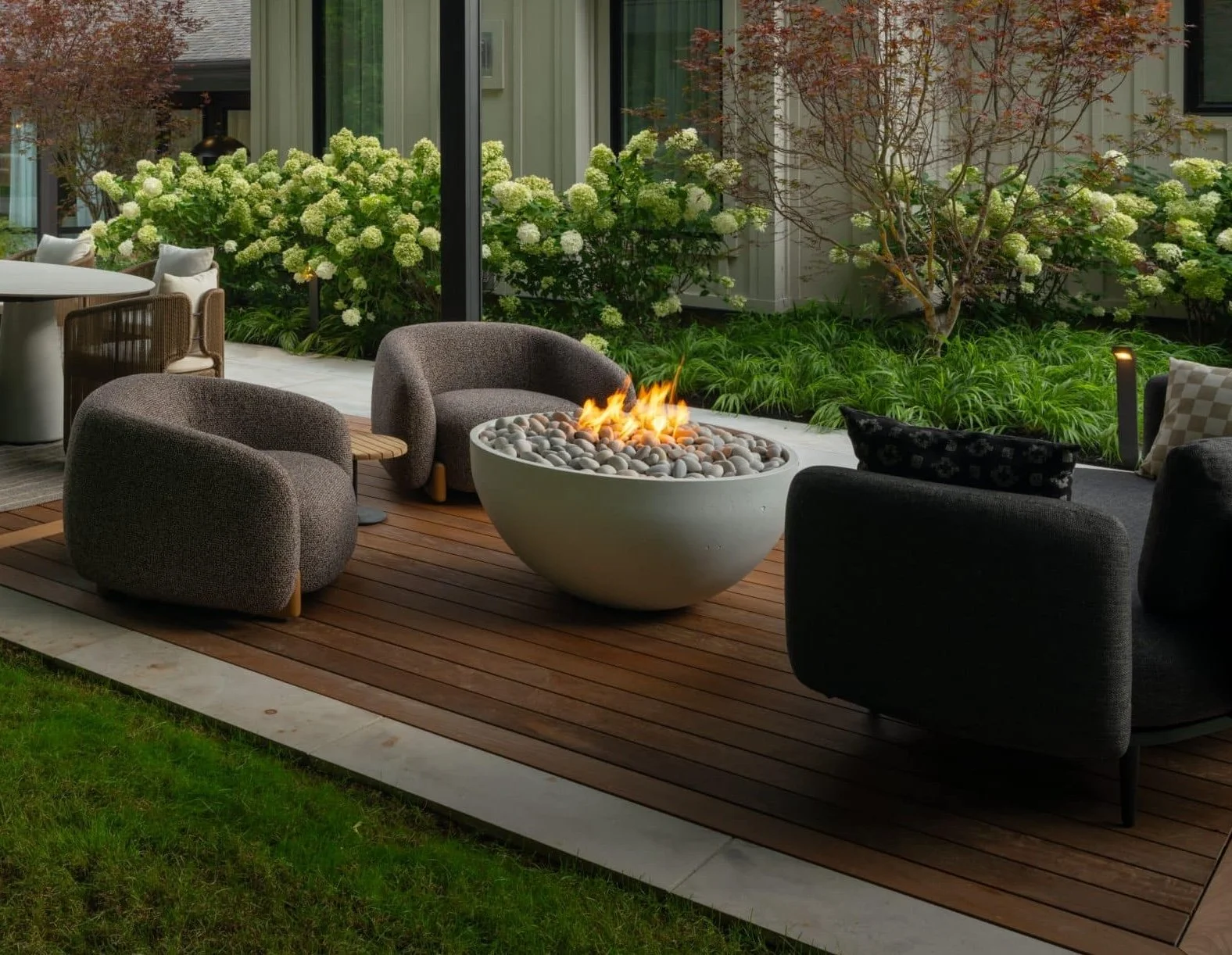 Custom fire pit set within an outdoor patio area with modern chairs and lush green plants. Part of a patio house with large glass windows.