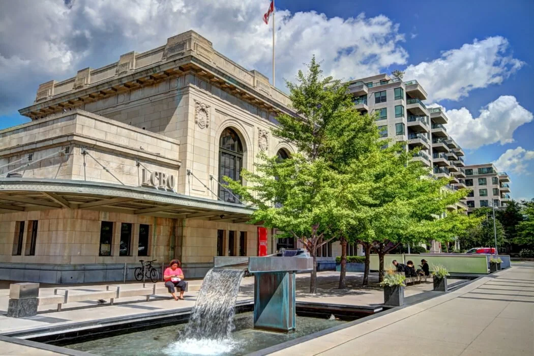 A historic building with the word 'LEBO' on the front, a water fountain in front, and several trees and benches along the sidewalk, with modern apartment buildings in the background.