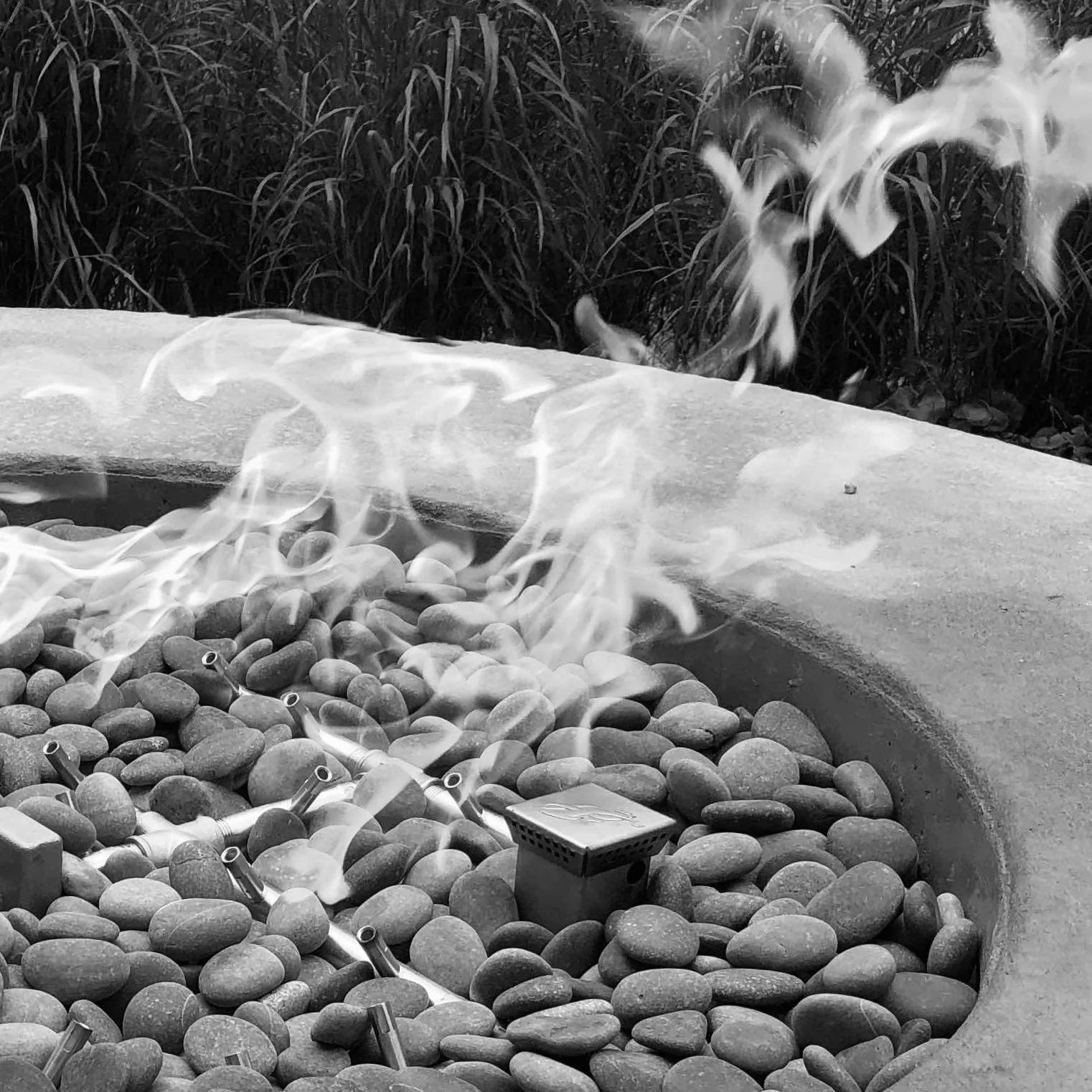Custom fire pit with flames and outdoor plants in the background.