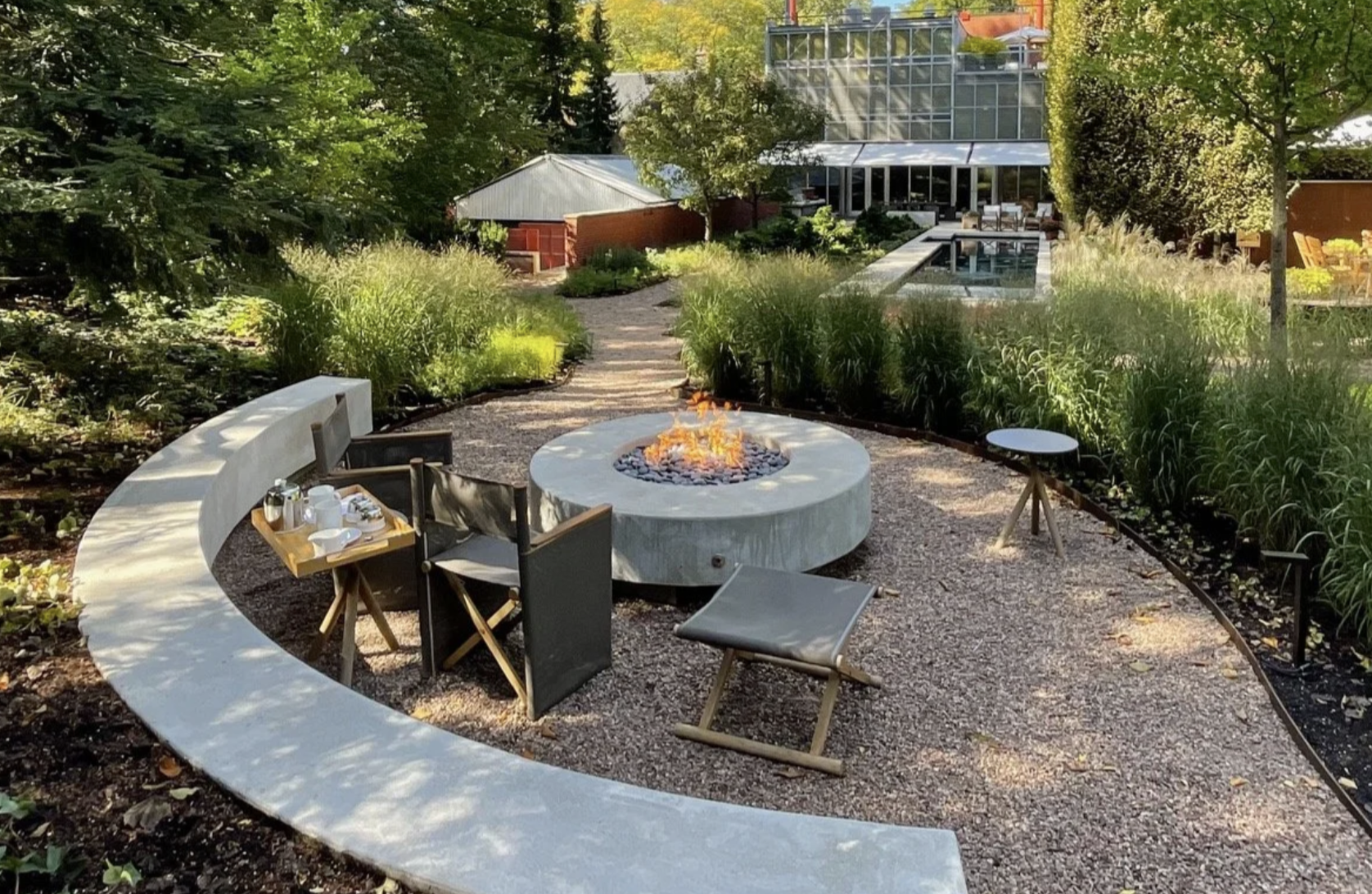 Outdoor garden seating area with a fire pit surrounded by chairs, a curved concrete bench, lush greenery, and a modern glass building in the background.