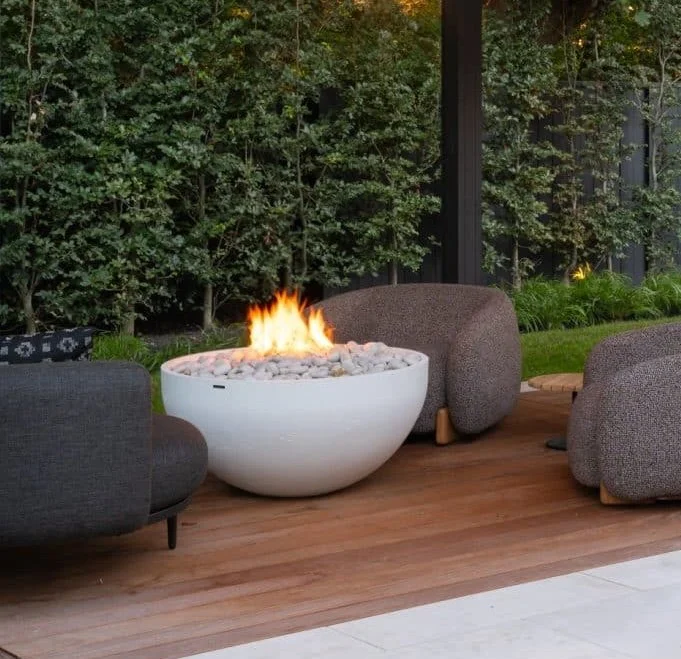 Custom fire pit set within an outdoor patio area with modern chairs and lush green plants. Part of a patio house with large glass windows.