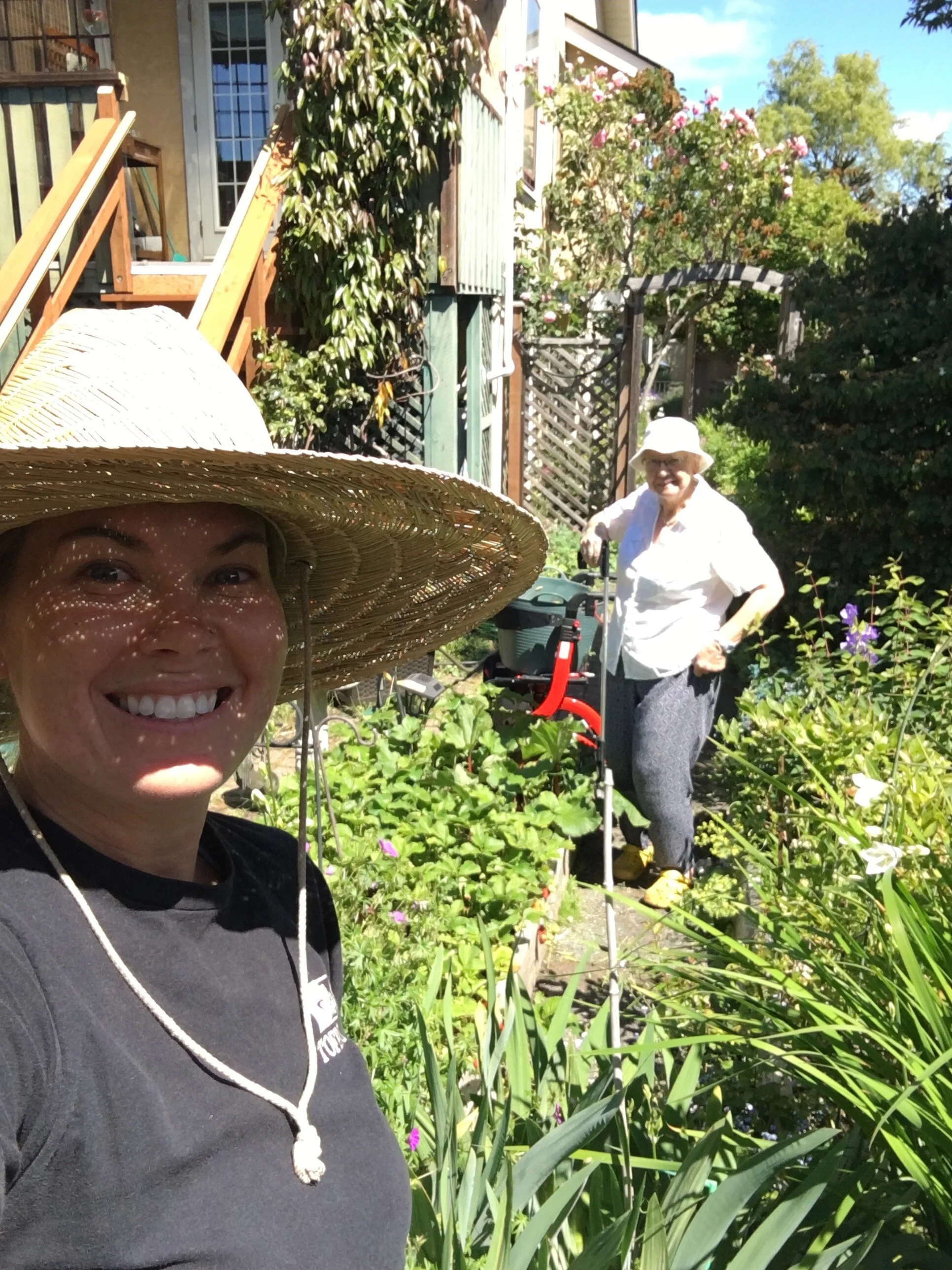 Two women in a garden with lush green plants, flowers, and a house in the background. One woman in the foreground is smiling and wearing a large straw hat, and the other woman in the background is standing near gardening tools.