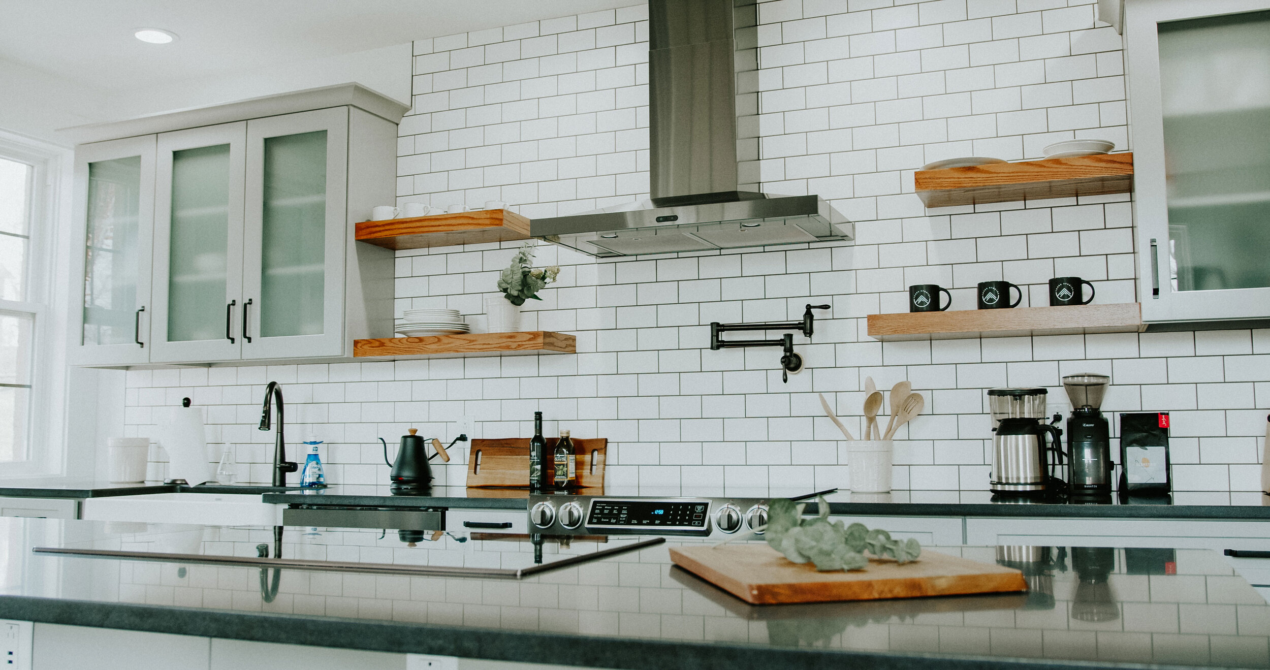 Modern kitchen with white subway tile backsplash, gray cabinets with frosted glass upper doors, wooden open shelves, black countertop, black kitchen faucet, stove, coffee maker, kitchen utensils, and decorative items.