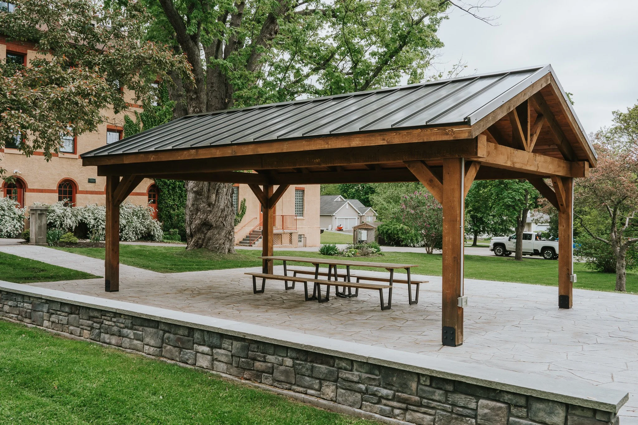 A wooden picnic shelter with a metal roof in a park, featuring a picnic table underneath, surrounded by trees, grass, and nearby houses.