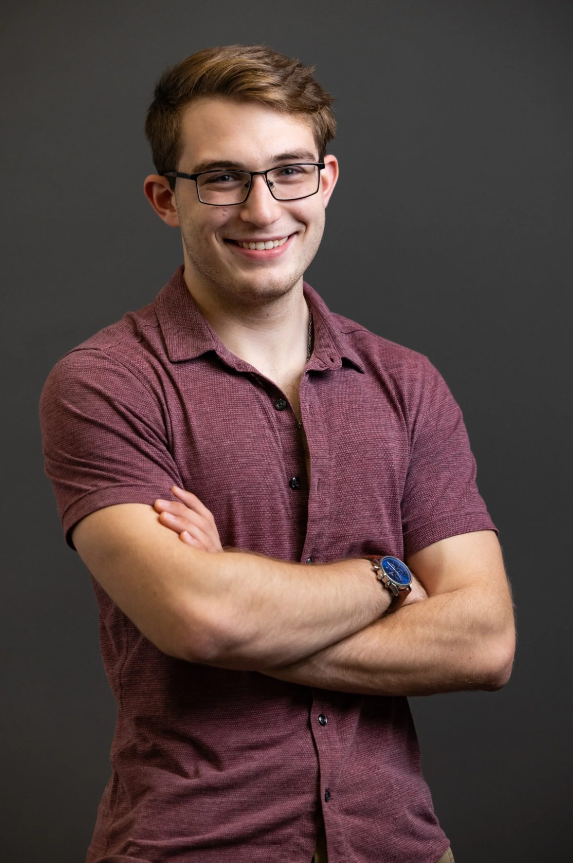 Young man with glasses smiling, arms crossed, wearing a maroon short-sleeve button-up shirt, dark background.