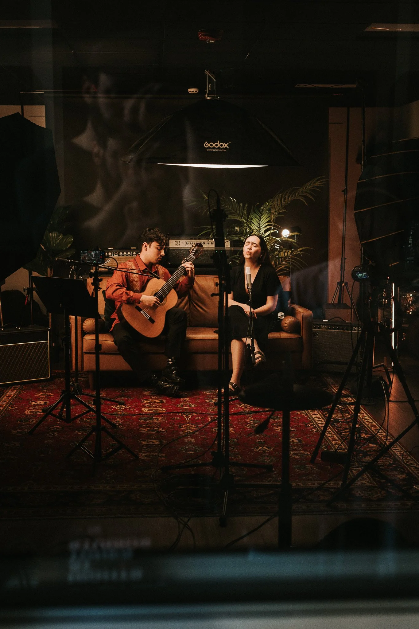 Two musicians, a man playing guitar and a woman singing, perform on a cozy stage with warm lighting, surrounded by recording equipment and large plants.