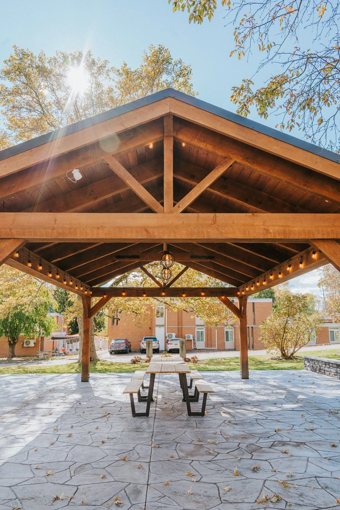 Wooden gazebo with string lights, a picnic table underneath, set in a park with trees and a brick building in the background, during daytime with sunlight.
