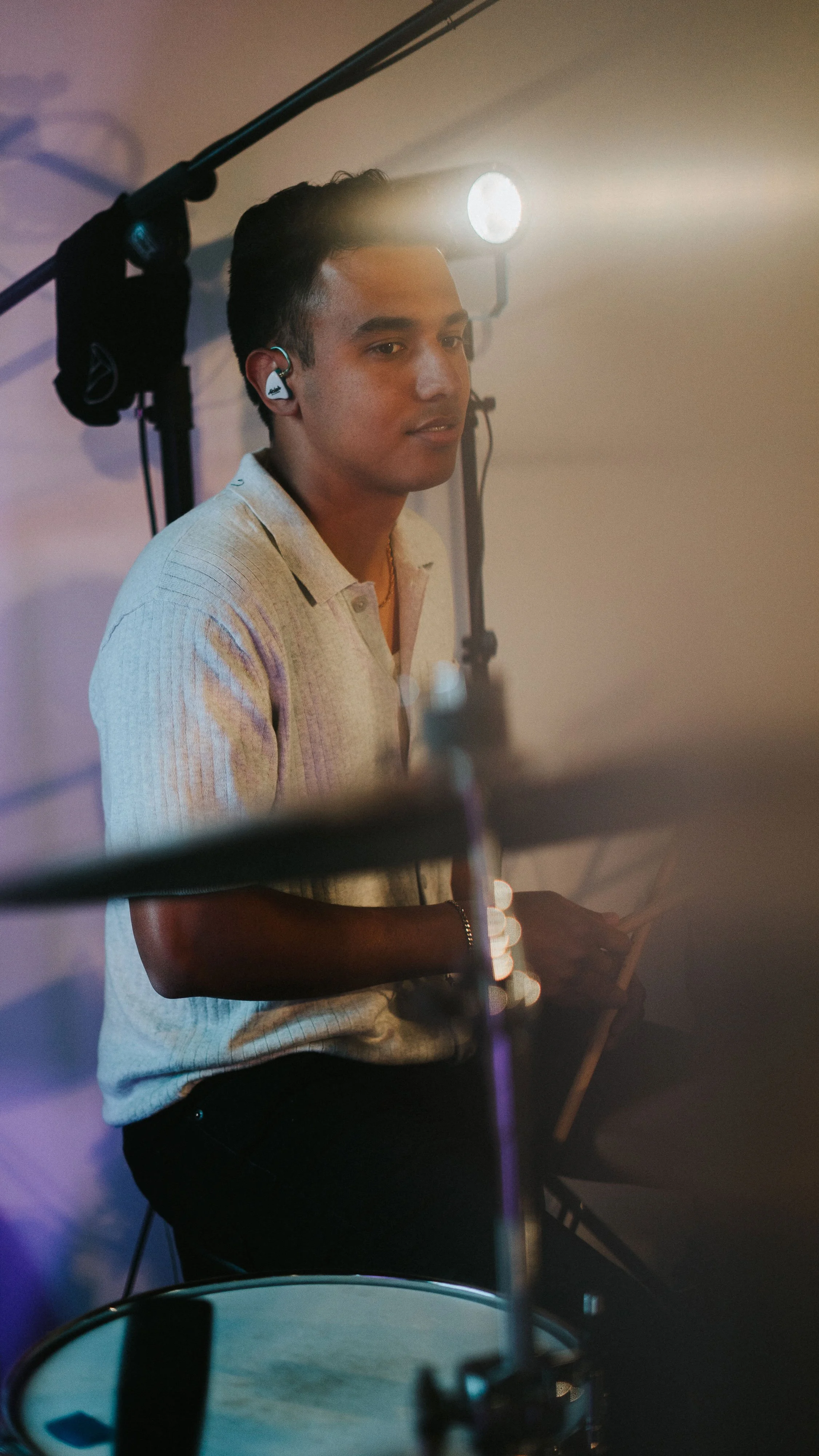 A young man with dark hair wearing a beige polo shirt and wireless earbuds, standing behind a drum set with stage lighting overhead.