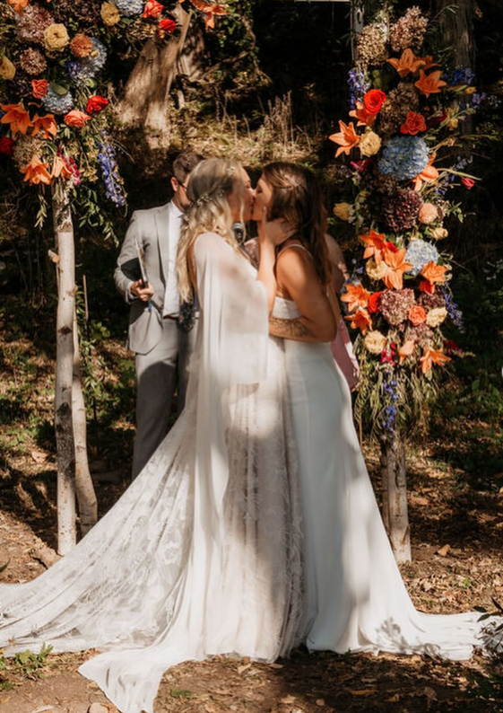 Couple getting married at an outdoor wedding ceremony, kissing under a floral arch with a officiant standing beside them, surrounded by trees.