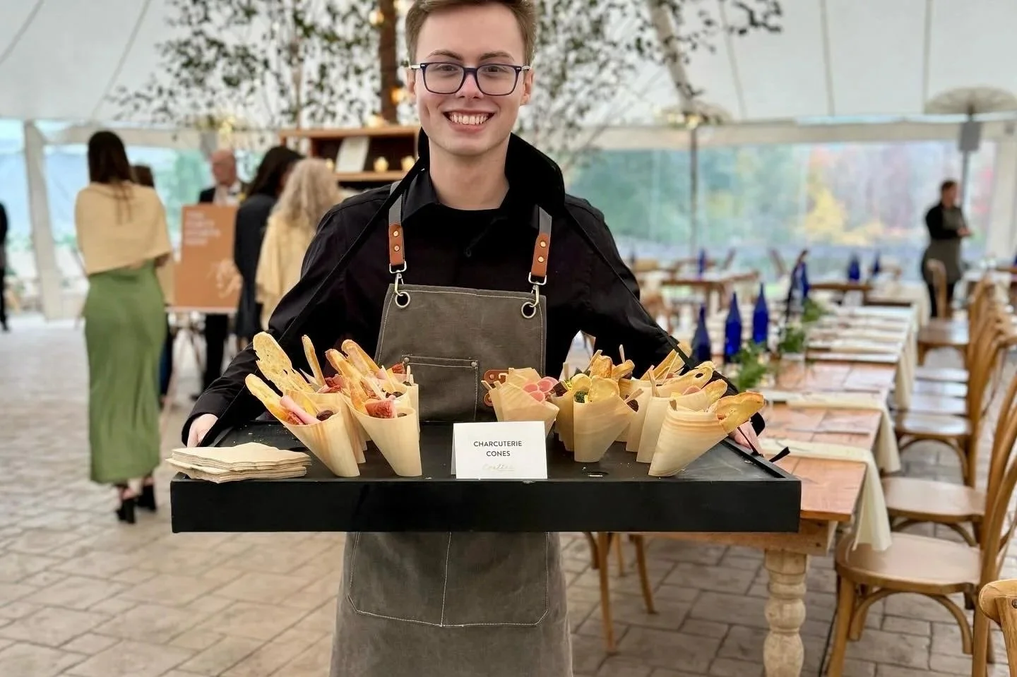 A smiling waiter in a black shirt and gray apron carrying a tray of charcuterie cones at a restaurant with tables and people in the background.