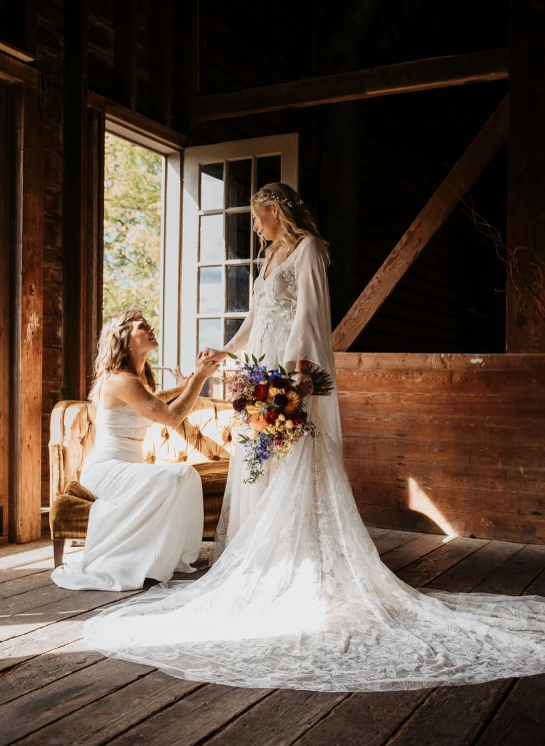 Two women, one in a wedding dress and the other in a white gown, in a rustic wooden room beside an open window with sunlight streaming in. The standing woman holds a bouquet of colorful flowers, and the seated woman is reaching out to her, creating a