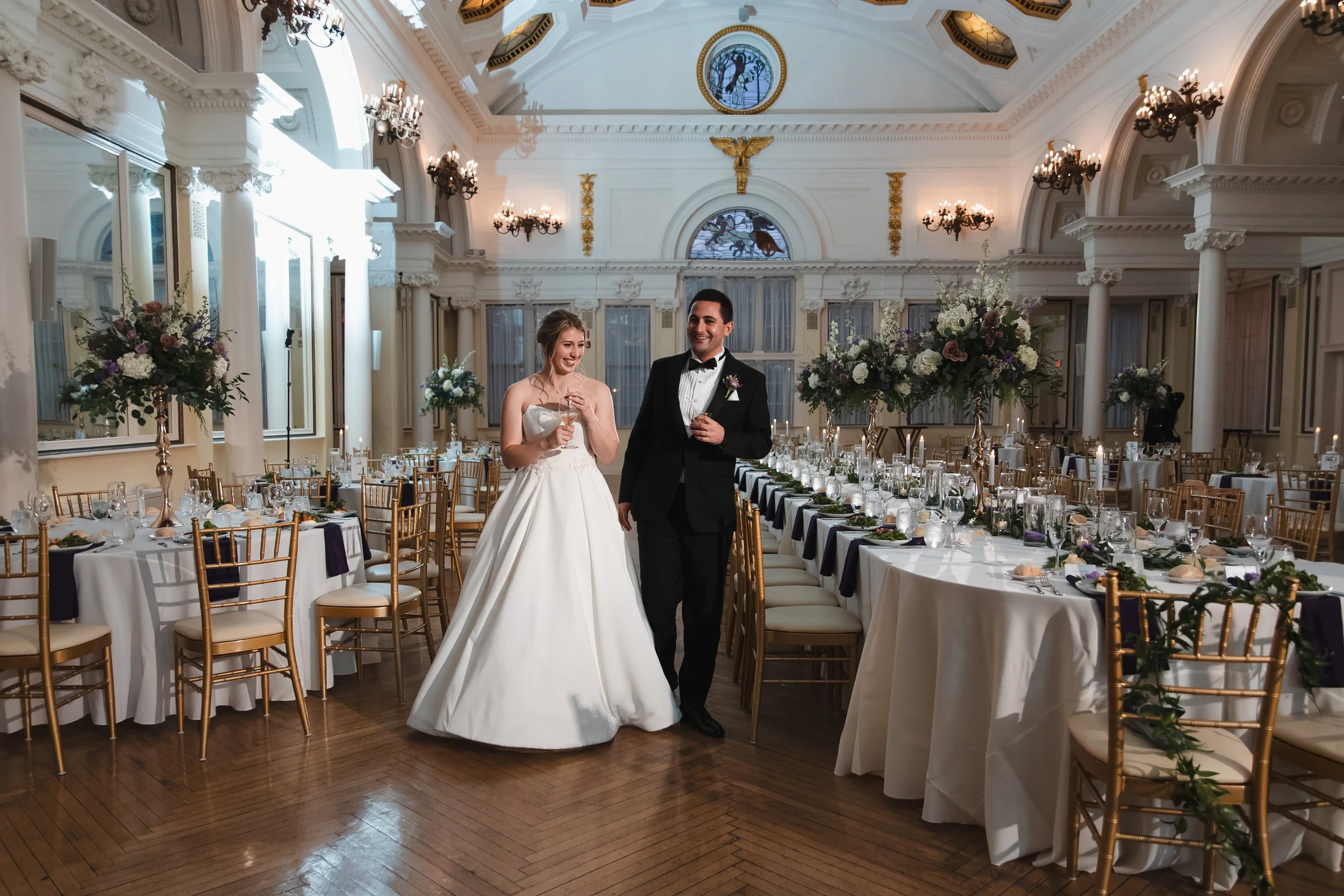 A bride and groom walking inside a decorated wedding reception hall, with the bride in a white wedding dress holding a glass, and the groom in a black tuxedo, surrounded by elegant tables with floral centerpieces and gold chairs.