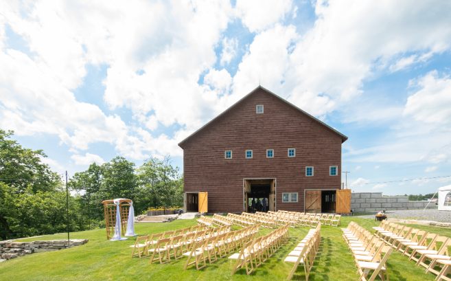 A rustic barn converted into a wedding venue with rows of wooden chairs set up on green grass, an outdoor altar with white drapes, and a cloudy blue sky.