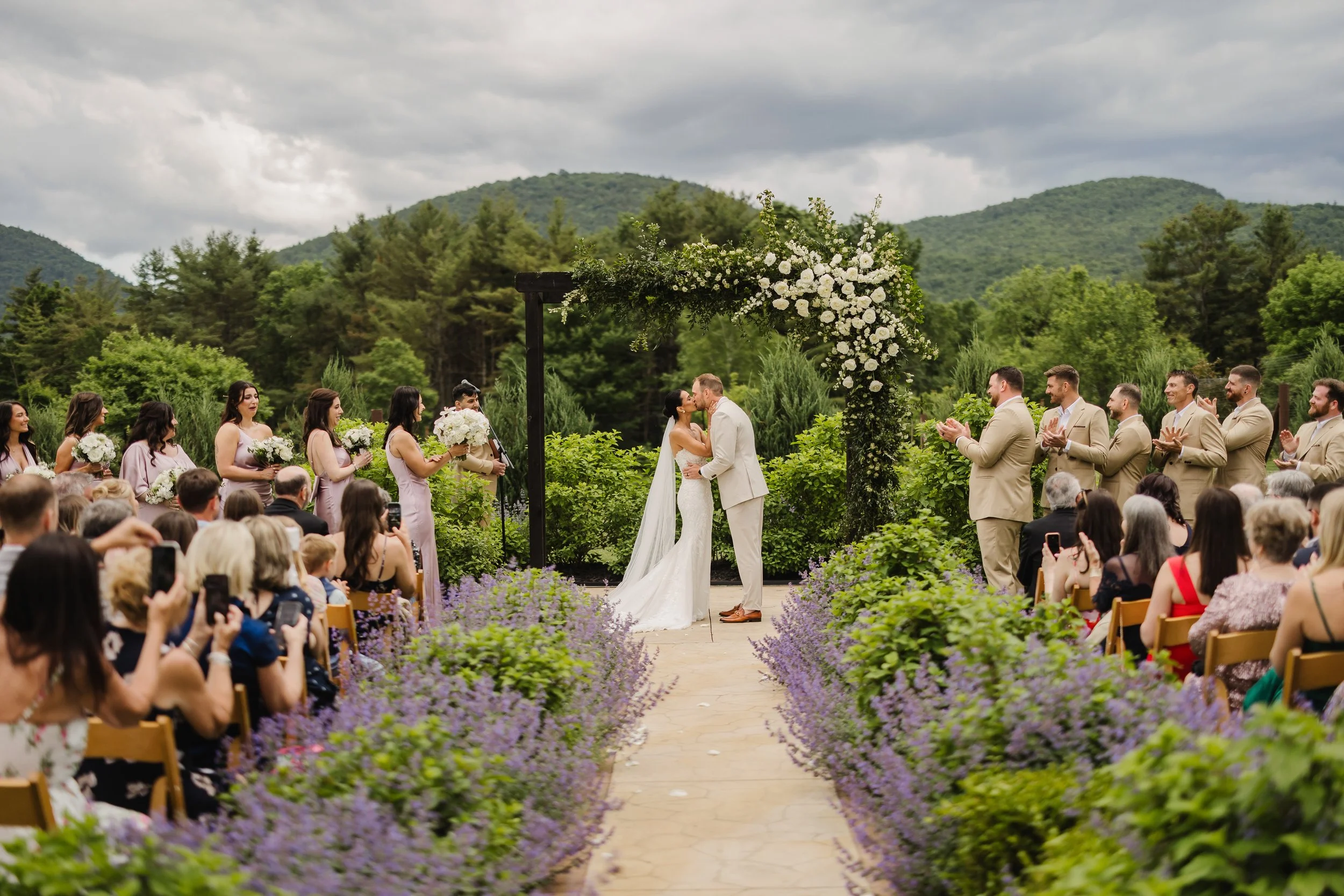 A couple kissing at their outdoor wedding ceremony surrounded by guests, purple flowers, and greenery with mountains in the background.