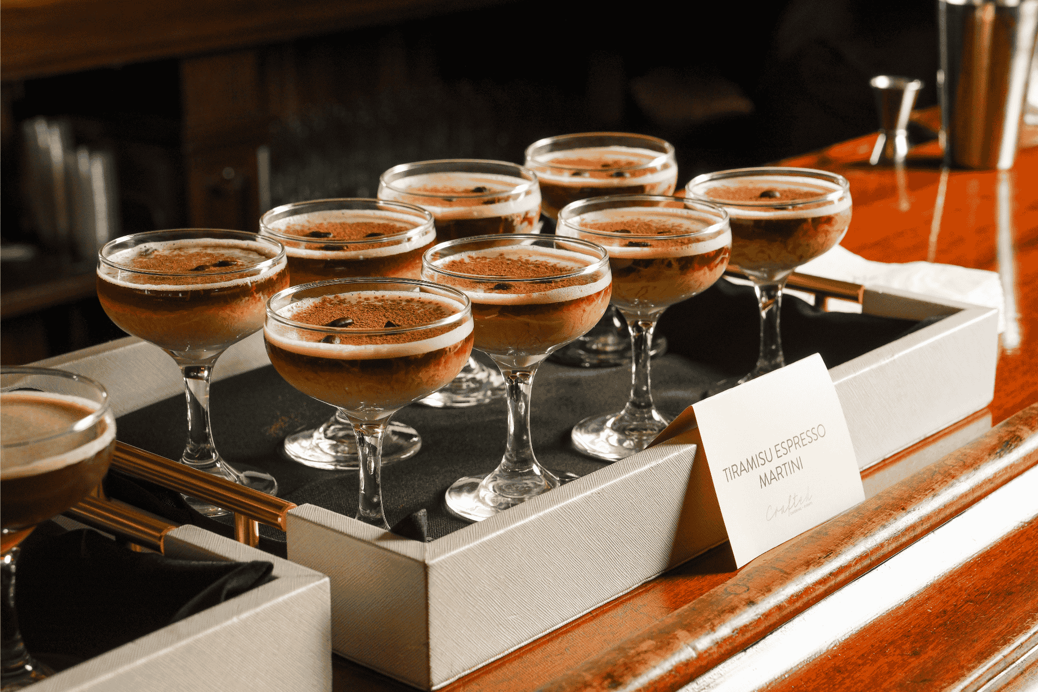 A tray of eight glasses filled with tiramisu espresso martini desserts on a wooden bar counter.