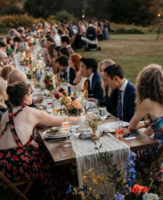 A large outdoor dinner party with many people seated long tables decorated with flowers and candles in a park or garden setting during sunset.