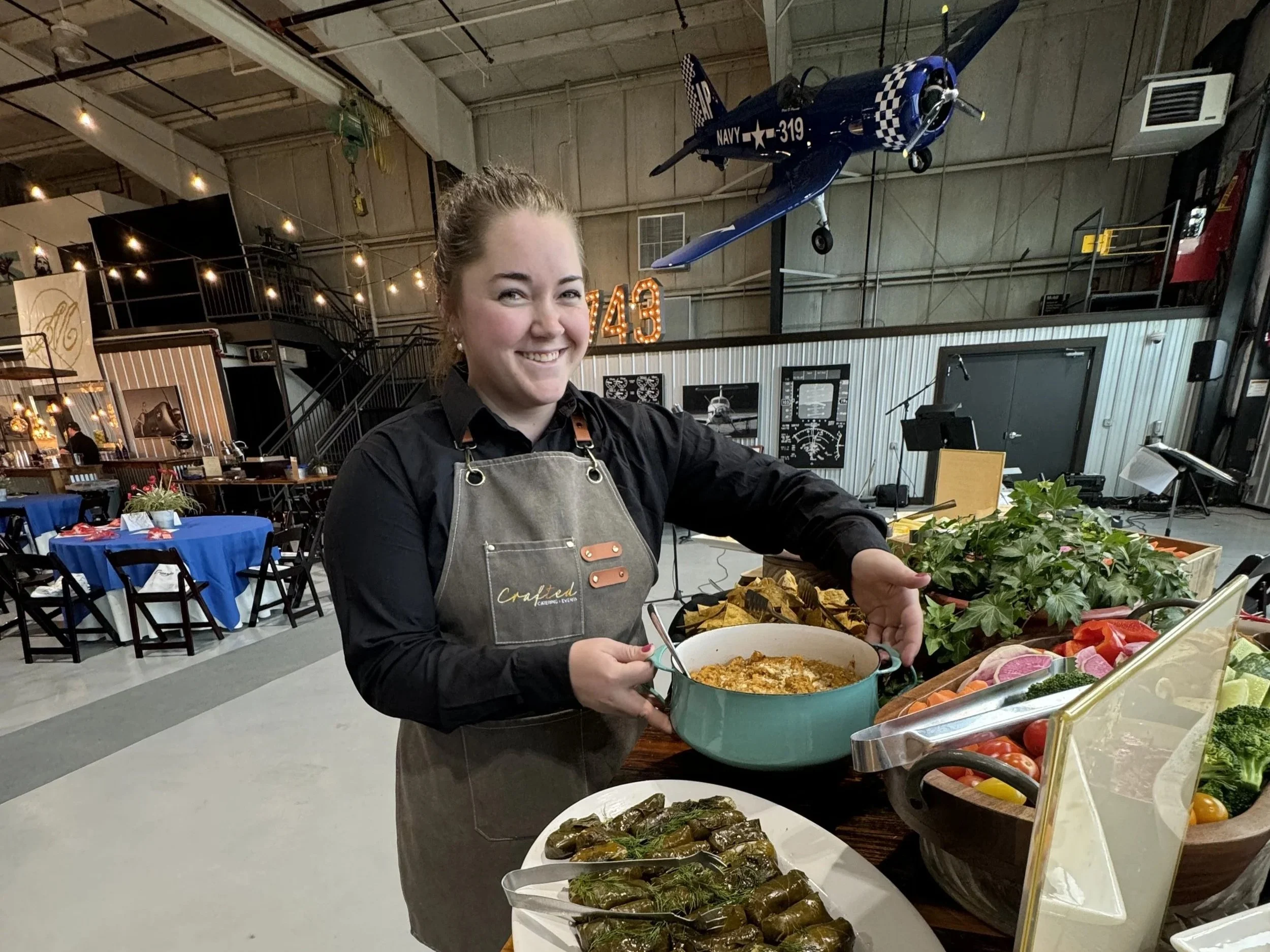 A young woman in an apron smiling and holding a dish at a farm-to-table event. Behind her are tables with blue tablecloths and a vintage airplane hanging from the ceiling in an industrial space.