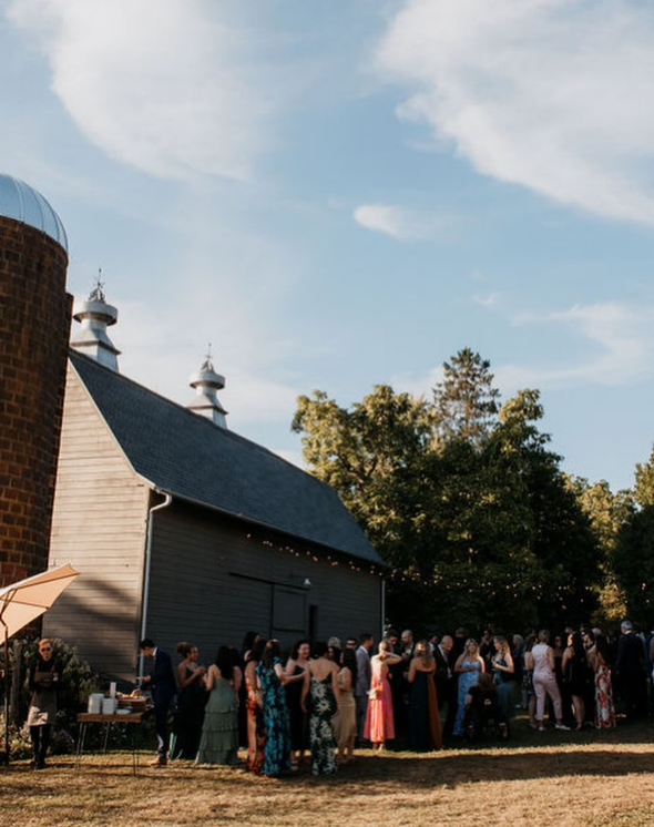 Group of people dressed in formal attire gathered outdoors near a rustic building with a large barn and silo, under a partly cloudy sky.