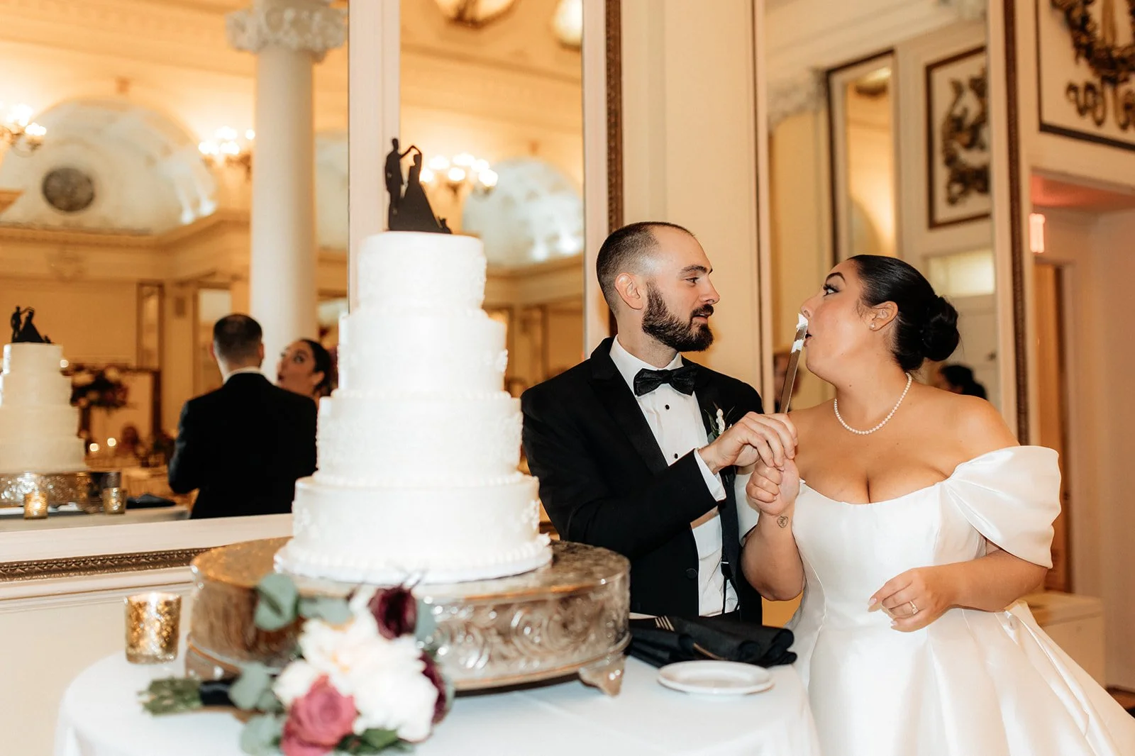 A bride and groom feeding each other wedding cake in a decorated reception hall, with a tiered wedding cake and floral arrangement on the table in front of them.