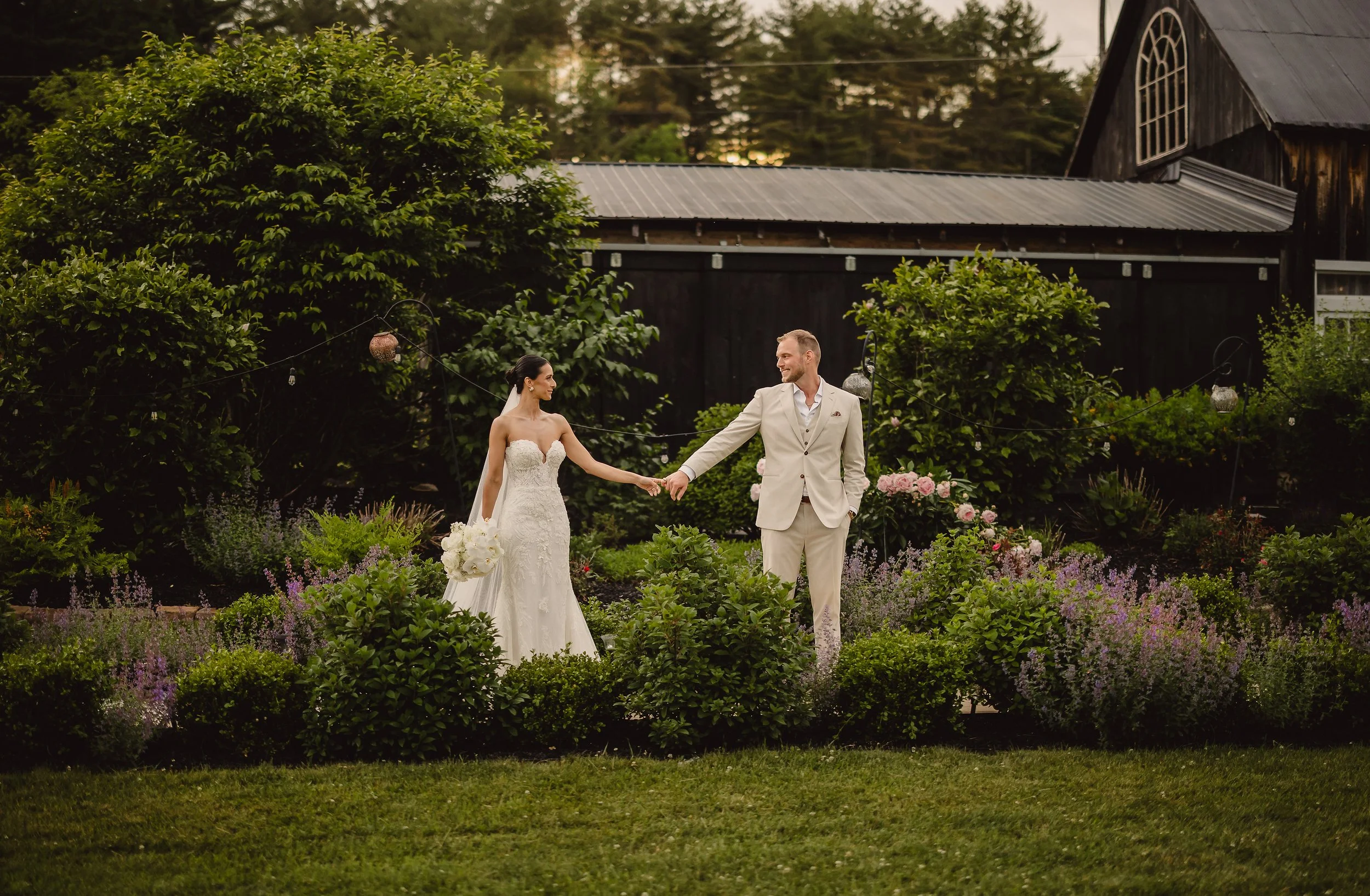 A bride and groom holding hands in a garden with flowers, bushes, and trees, during sunset.