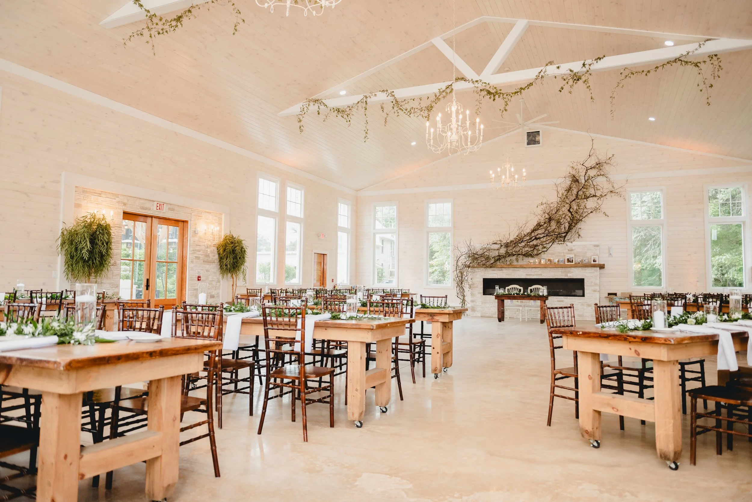 Bright, airy event hall with high wooden ceiling, large windows, and elegant light fixtures. There are wooden tables and chairs arranged for a gathering, with greenery and white wreaths as table decorations. The fireplace has a branch with vines exte