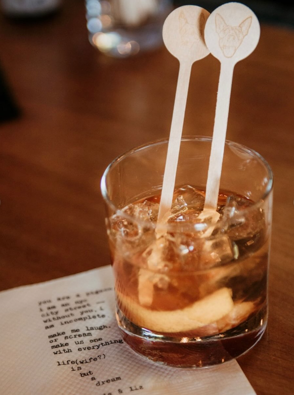 A glass of iced beverage with two drink stirrers, one with a cat's face drawing, placed on a wooden table next to a piece of paper with typed text.