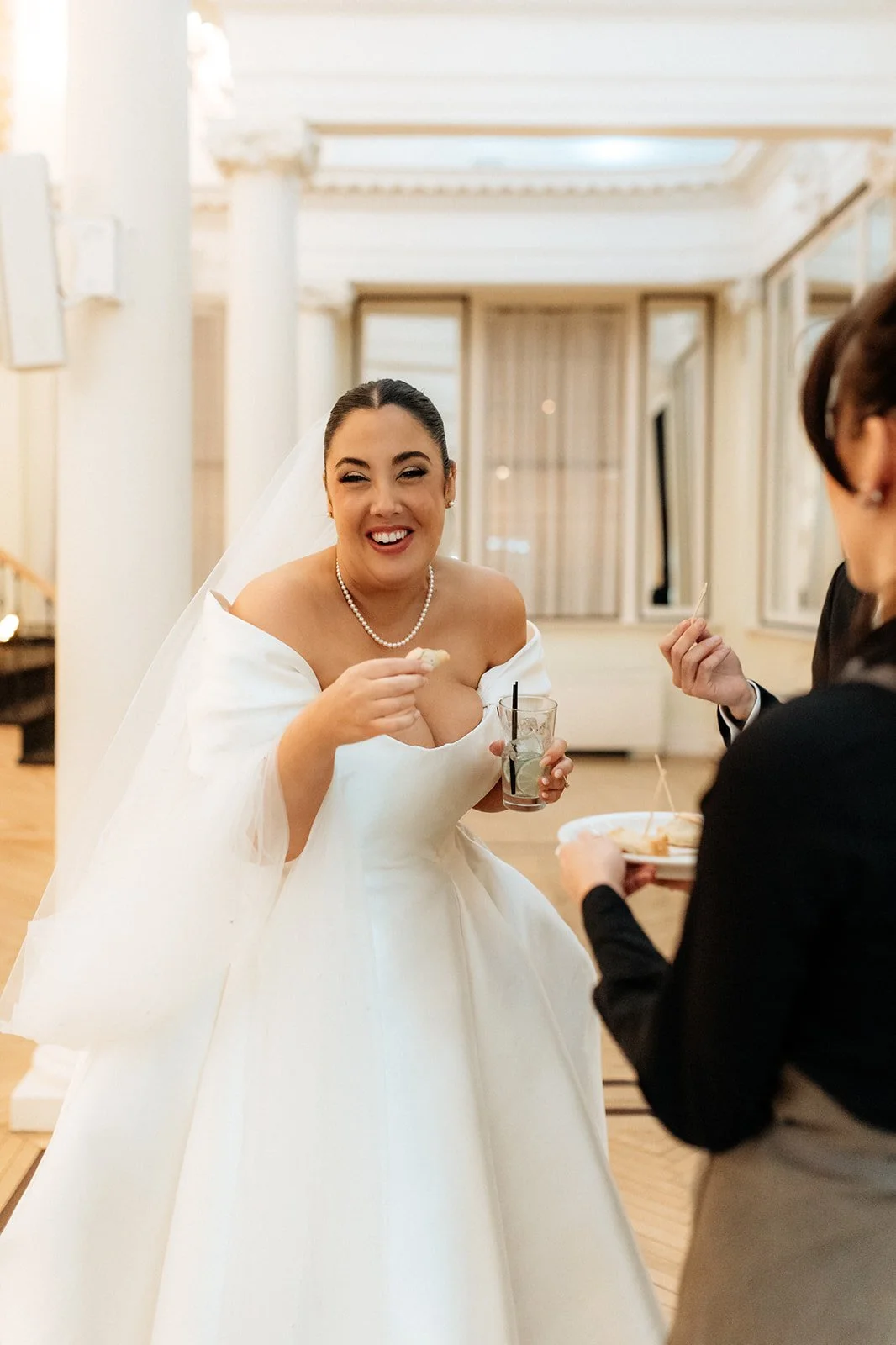 Bride in a white wedding gown and veil smiling while holding a drink and a cookie, talking to a person in a black blazer, at a wedding reception indoors.