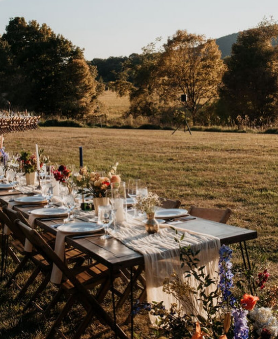 Long outdoor dining table set for a celebration with floral centerpieces, surrounded by folding chairs, on a grassy field during sunset with trees in the background.