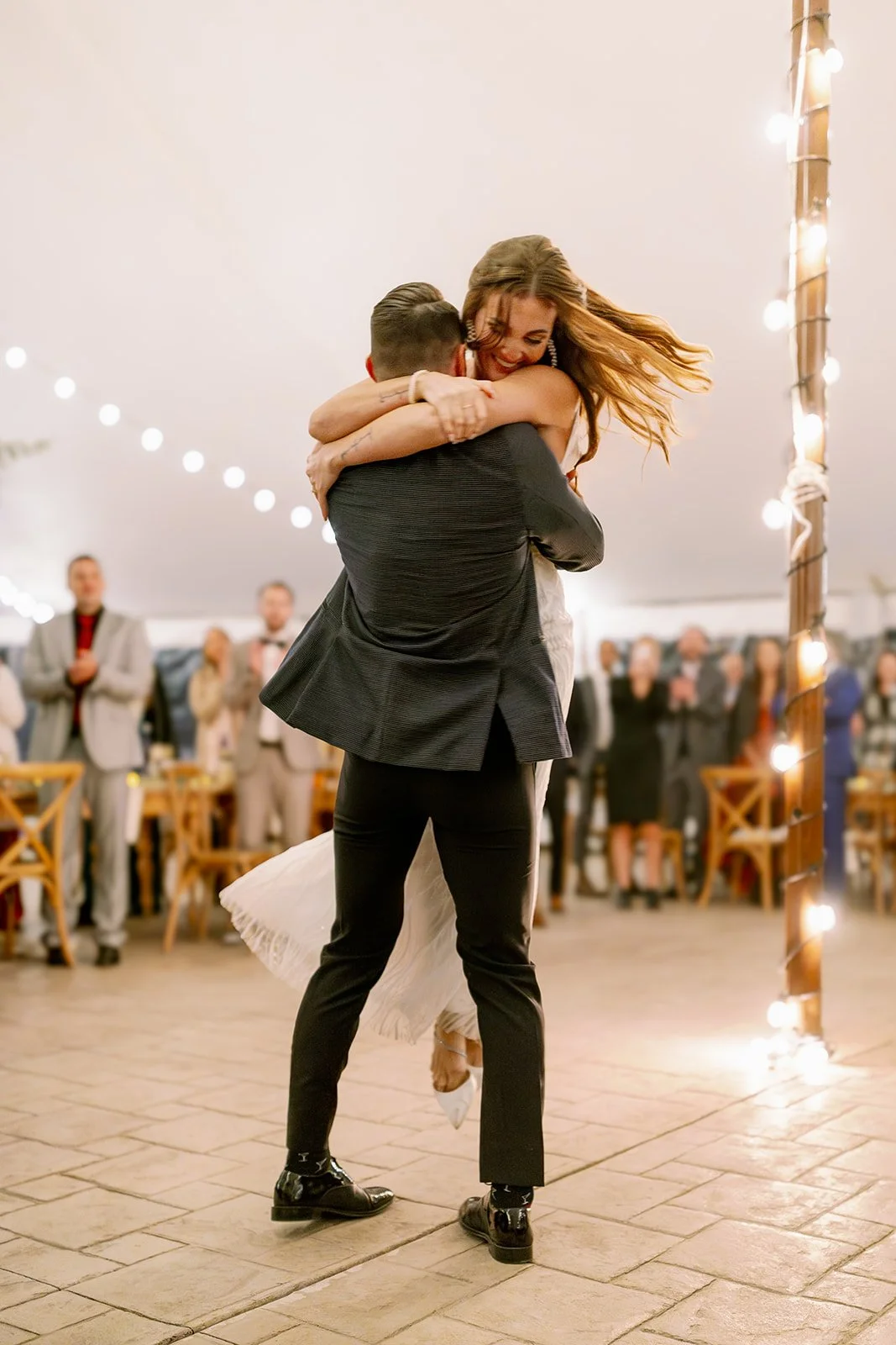 A couple dancing and hugging at a wedding reception, with guests watching in the background and string lights hanging from the ceiling.