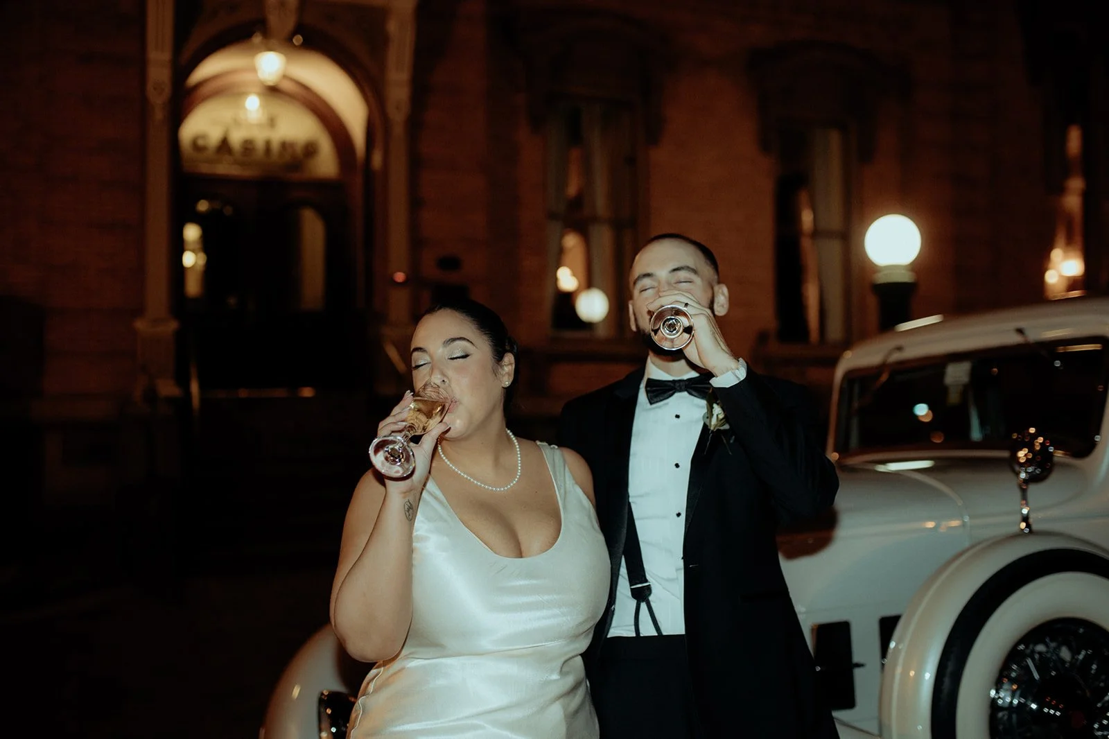 A bride and groom in wedding attire drinking champagne at night in front of a vintage car and illuminated building.