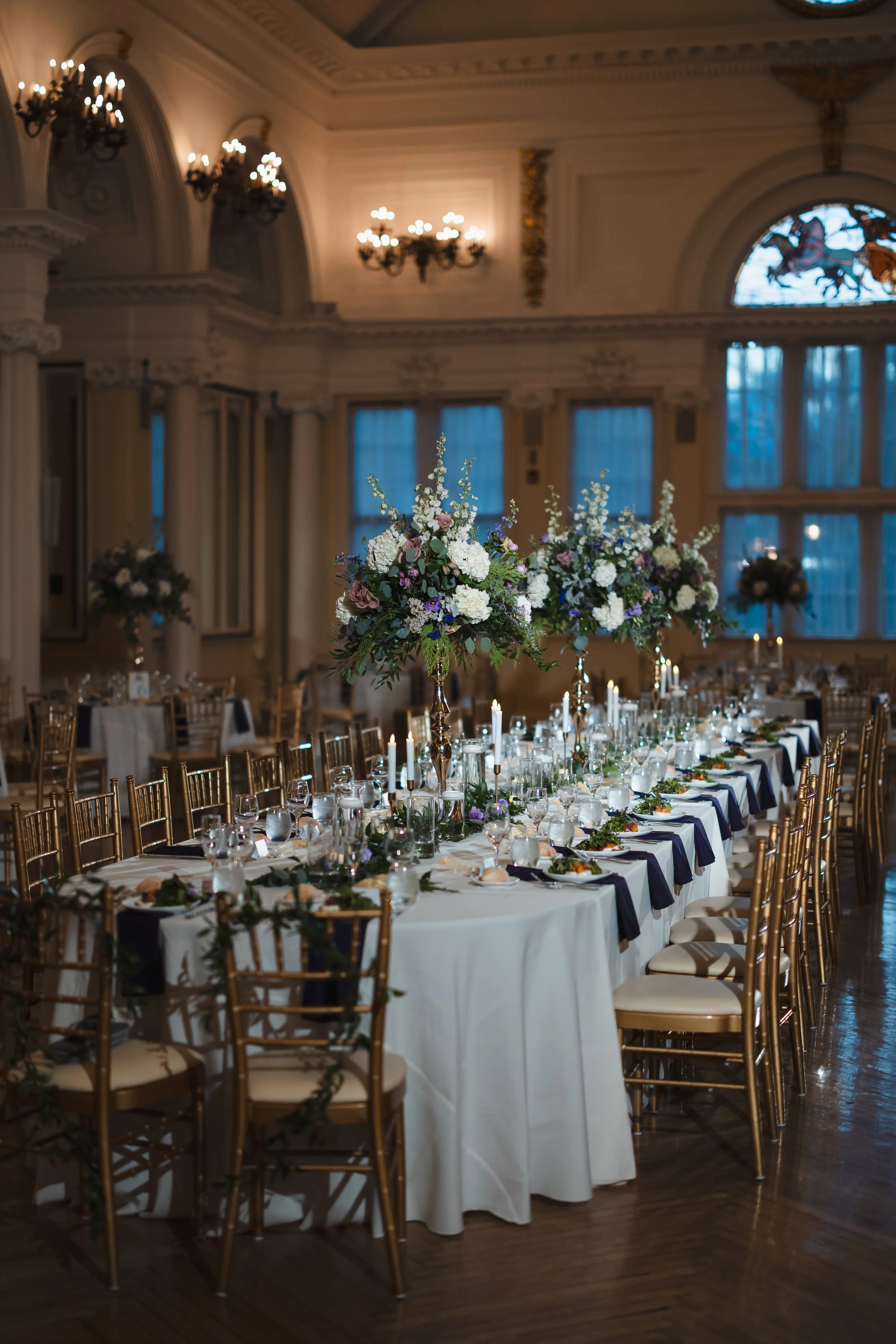 Elegant banquet table set for a wedding or formal event in an ornate, historic-style room with high ceilings, chandeliers, and large windows. The table features tall floral centerpieces with white, purple, and pink flowers, surrounded by lit candles,